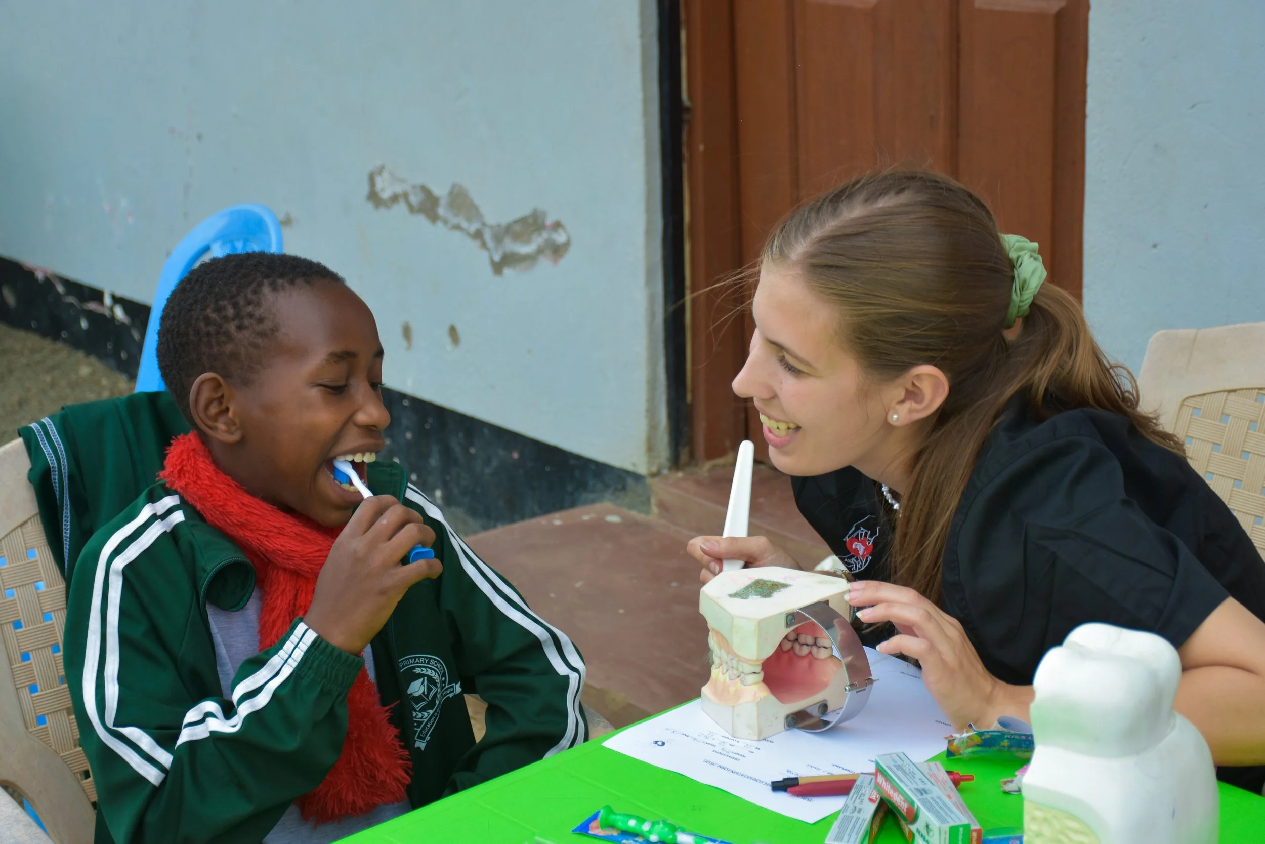A young orphan in a green jacket and red scarf sitting at a table, smiling and brushing his teeth with a toothbrush, while a smiling young woman dentist student with a ponytail holding a dental model and a toothbrush helping and teaching him.