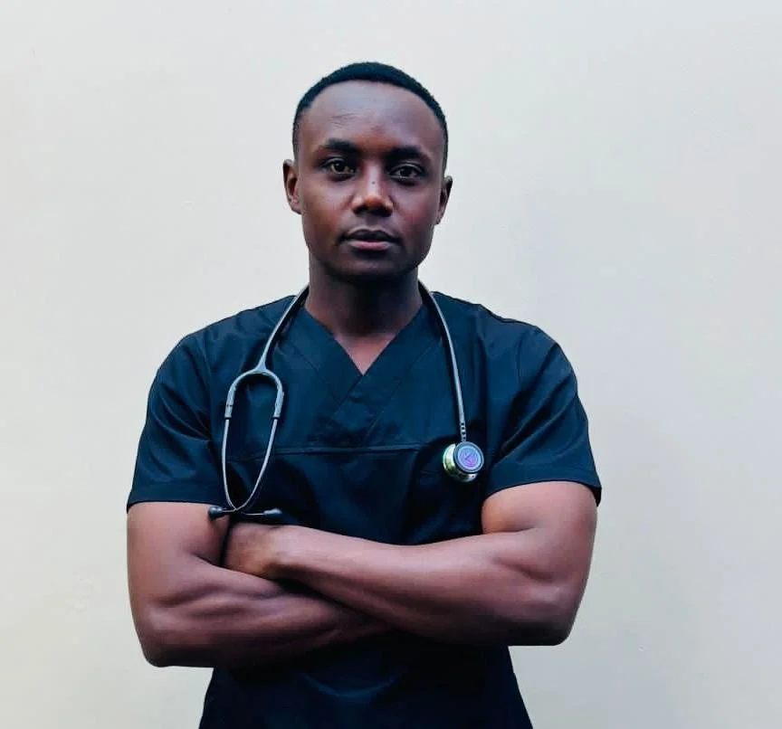 A male healthcare professional wearing dark scrubs and a stethoscope around his neck, standing against a plain background with arms crossed.