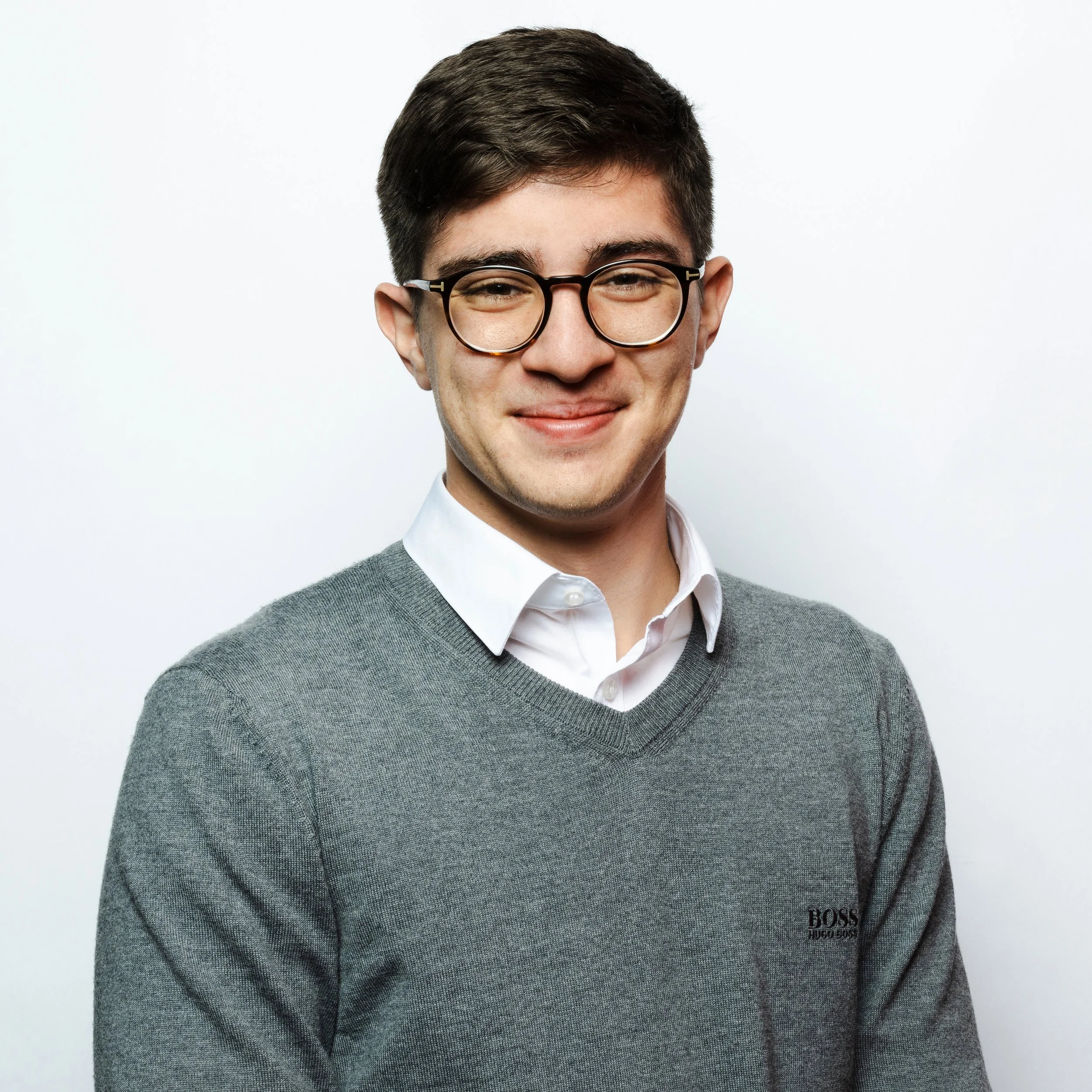 Young man wearing glasses, white collared shirt, and gray sweater with Hugo Boss logo, smiling against a plain white background.