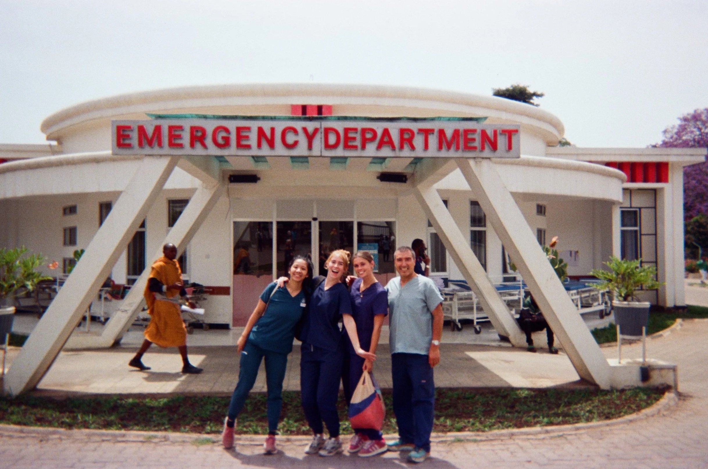 Four smiling volunteers in scrubs standing in front of an emergency department of Mount Meru Hospital Tanzania entrance, with a Maasai walking by in the background.