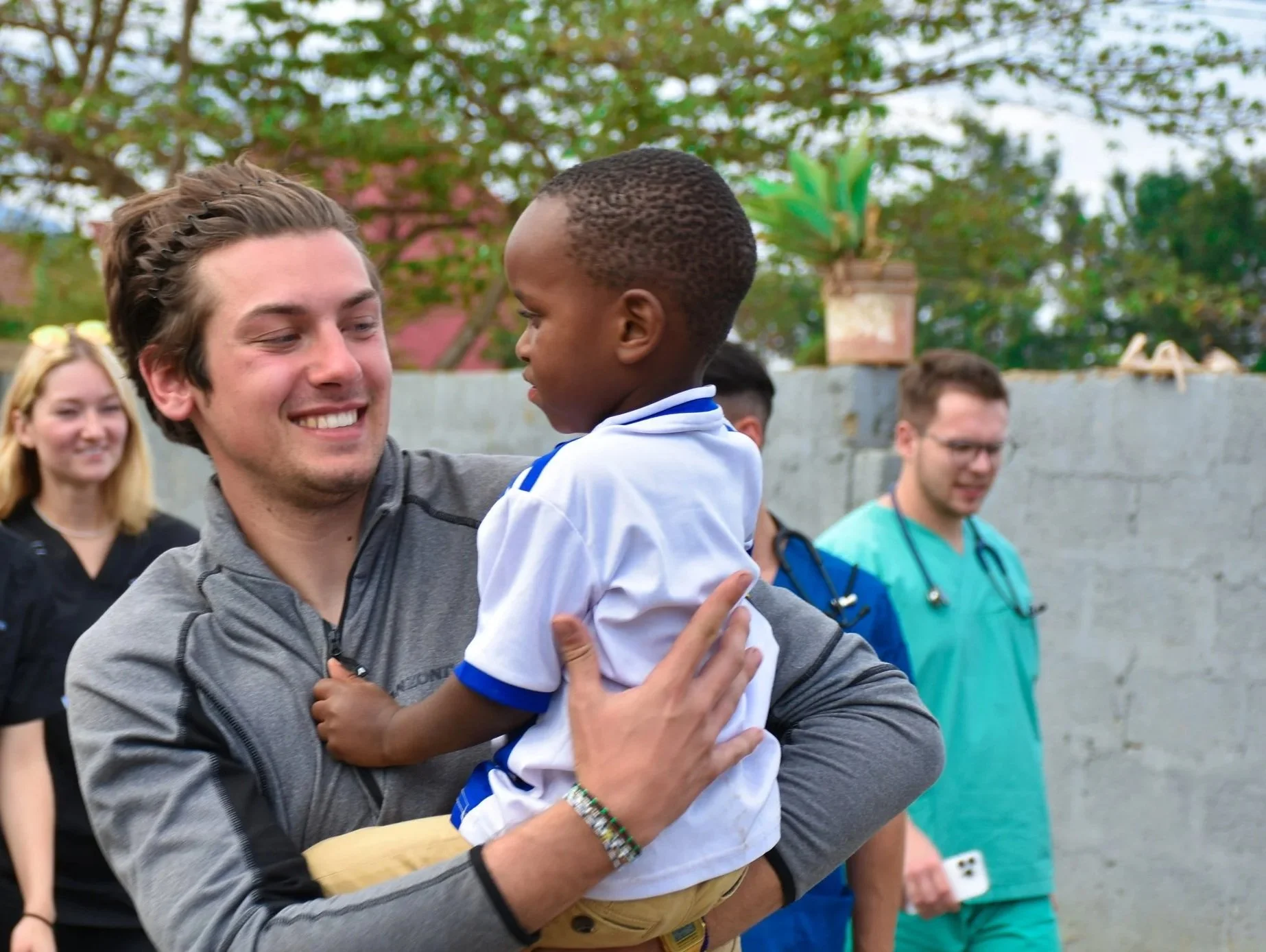A male volunteer at an orphanage holding an orphan child during a community outreach program. Behid stand students with stethoscopes