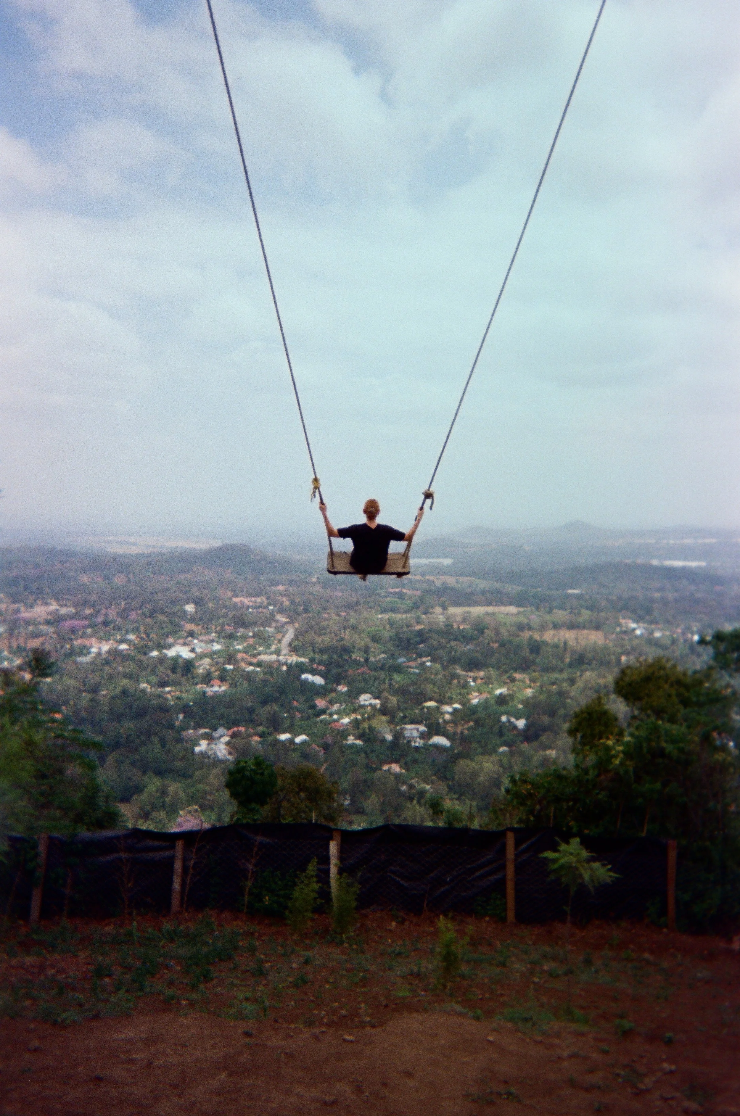 A person sitting on a swing hanging from two cables above a landscape with trees, houses, and distant hills under a cloudy sky.