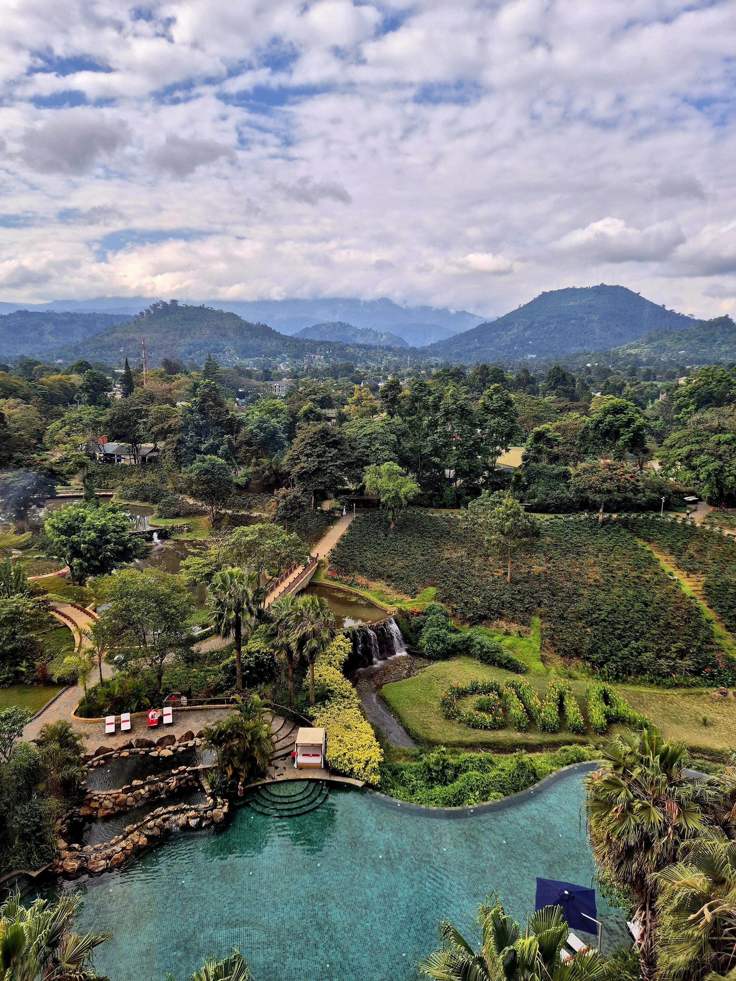 A scenic view of a lush green landscape with trees, mountains in the background, a small waterfall, a swimming pool, and a garden with the word 'LOVE' spelled out with flowers.