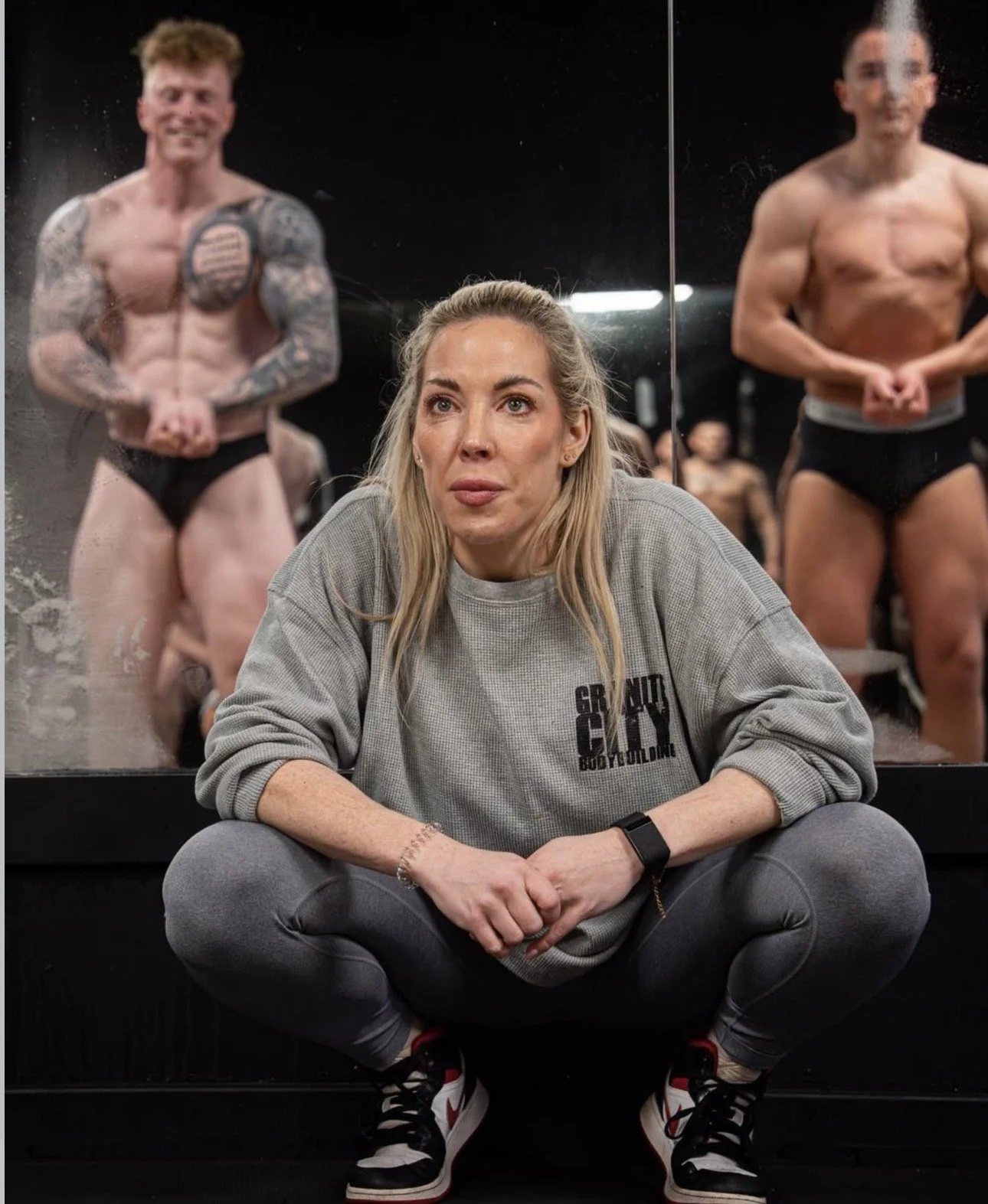 A posing coach sitting on the floor in front of a mirror with two bodybuilders learning bodybuilding posing for PCA, WNBF UK, UKDFBA