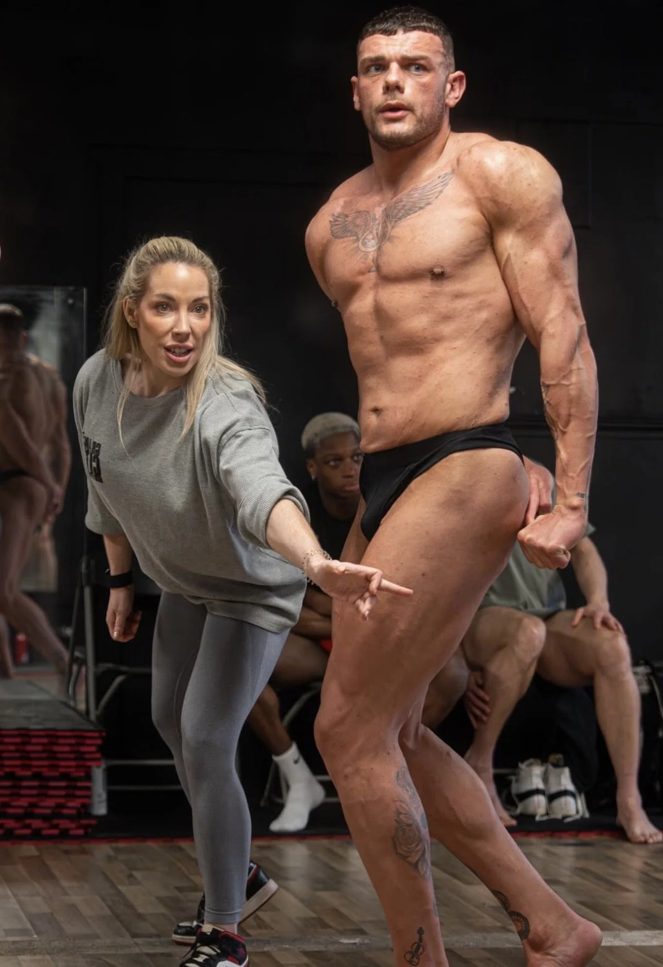 A posing coach points at a first timer bodybuilder, who is flexing his muscles to learn posing to go on stage. PCA, WNBF UK, UKDFBA