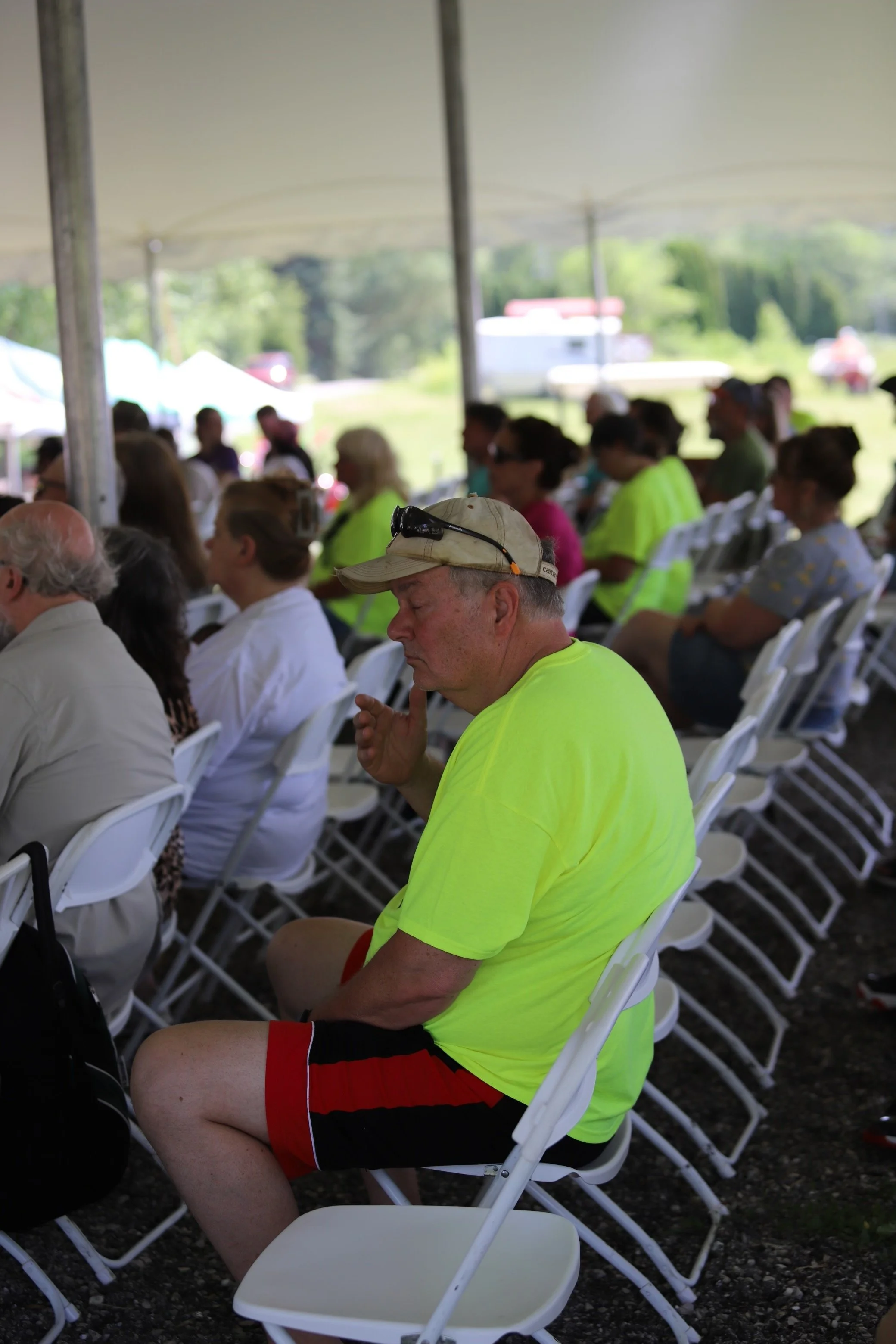 People sitting under a tent at a gathering, with some wearing bright yellow and neon clothing, and tall grass and trees in the background.
