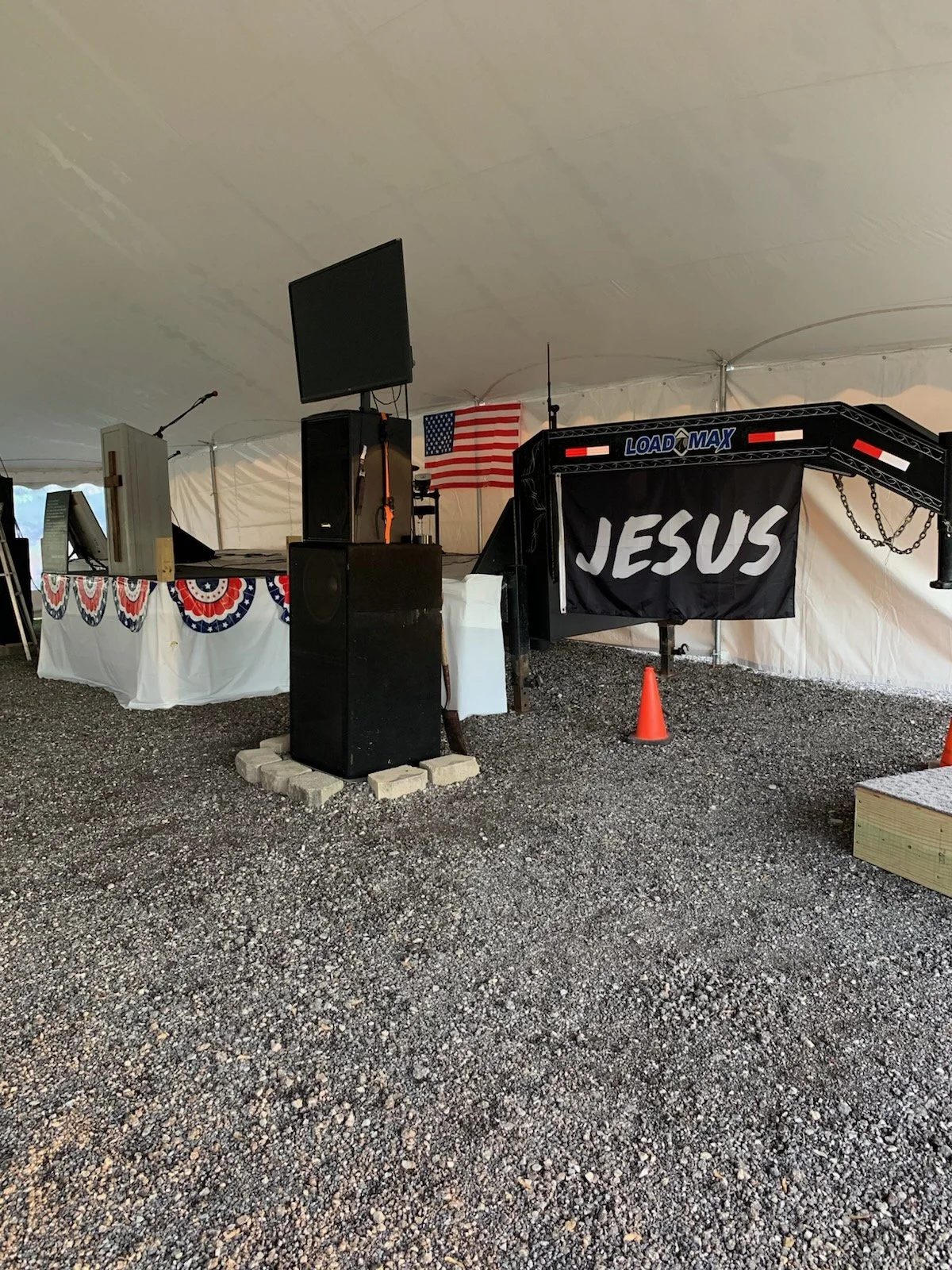 Inside a large event tent decorated with patriotic bunting, a stage with a black banner saying 'JESUS', American flags, and audio equipment, with gravel flooring and orange traffic cones.