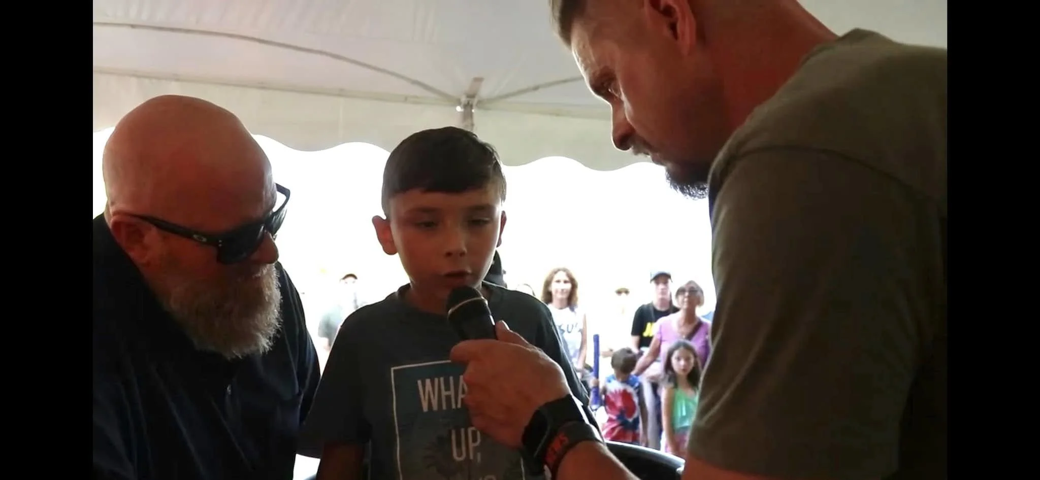 Three people, including a boy, an older man with glasses and a beard, and another man holding a microphone, are engaging in an activity under a tent at an event with a crowd of onlookers.