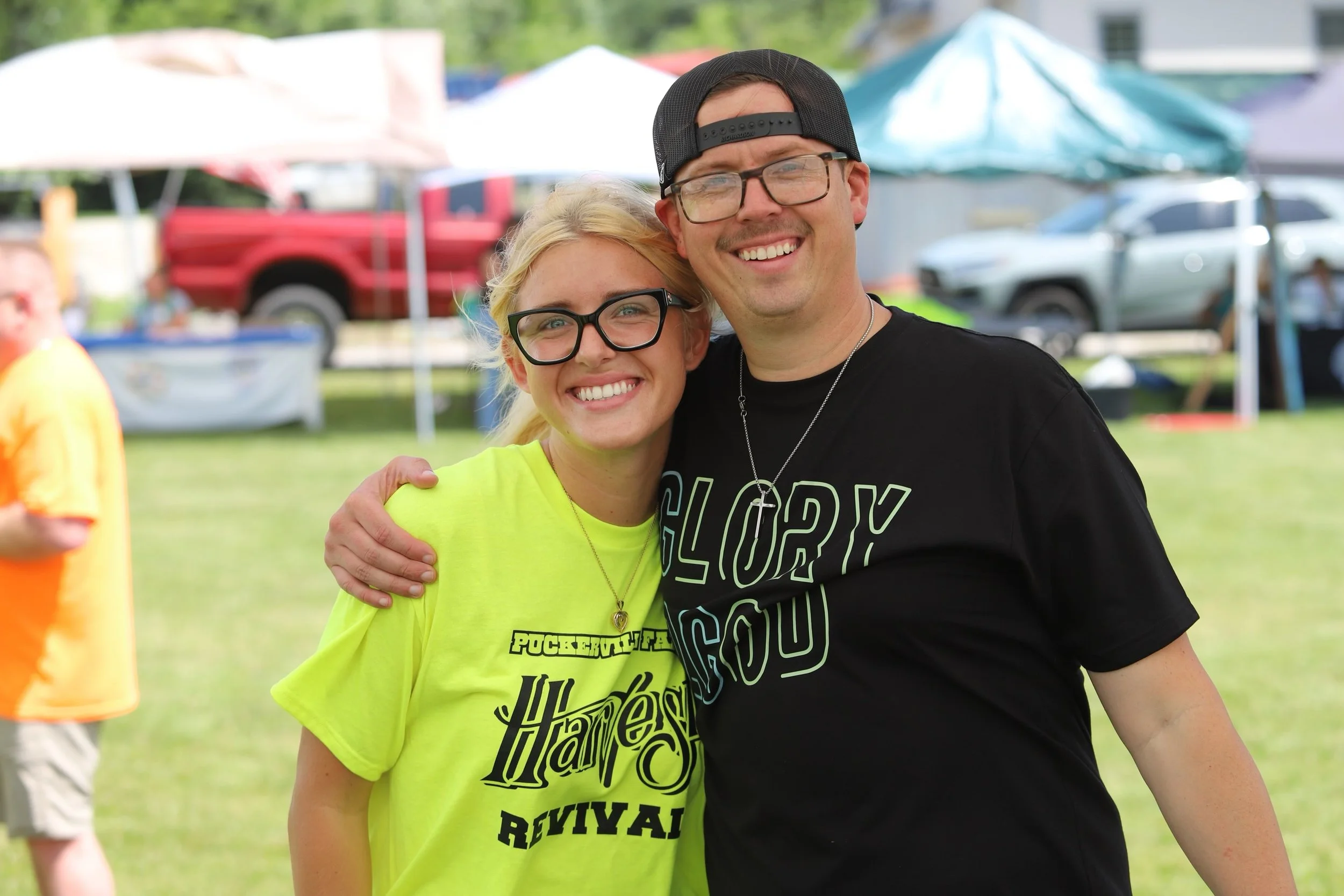 Two smiling people, a woman with blonde hair and glasses wearing a neon yellow shirt and a man with glasses and a black cap, standing outdoors at a festival or event.