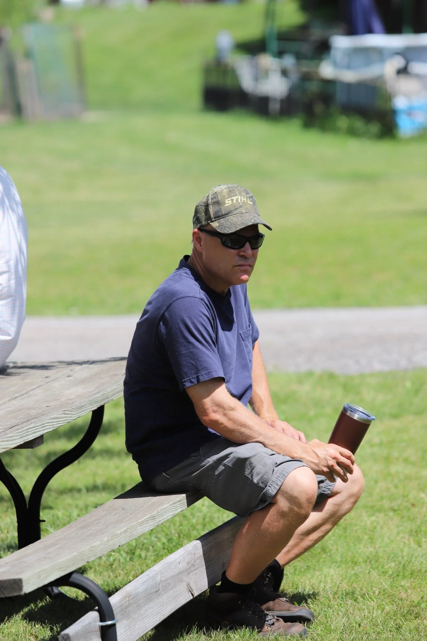Man sitting on a park bench, wearing sunglasses, a camouflage cap with the word 'STIHL', a navy T-shirt, gray shorts, and brown shoes, holding a tumbler with a lid.