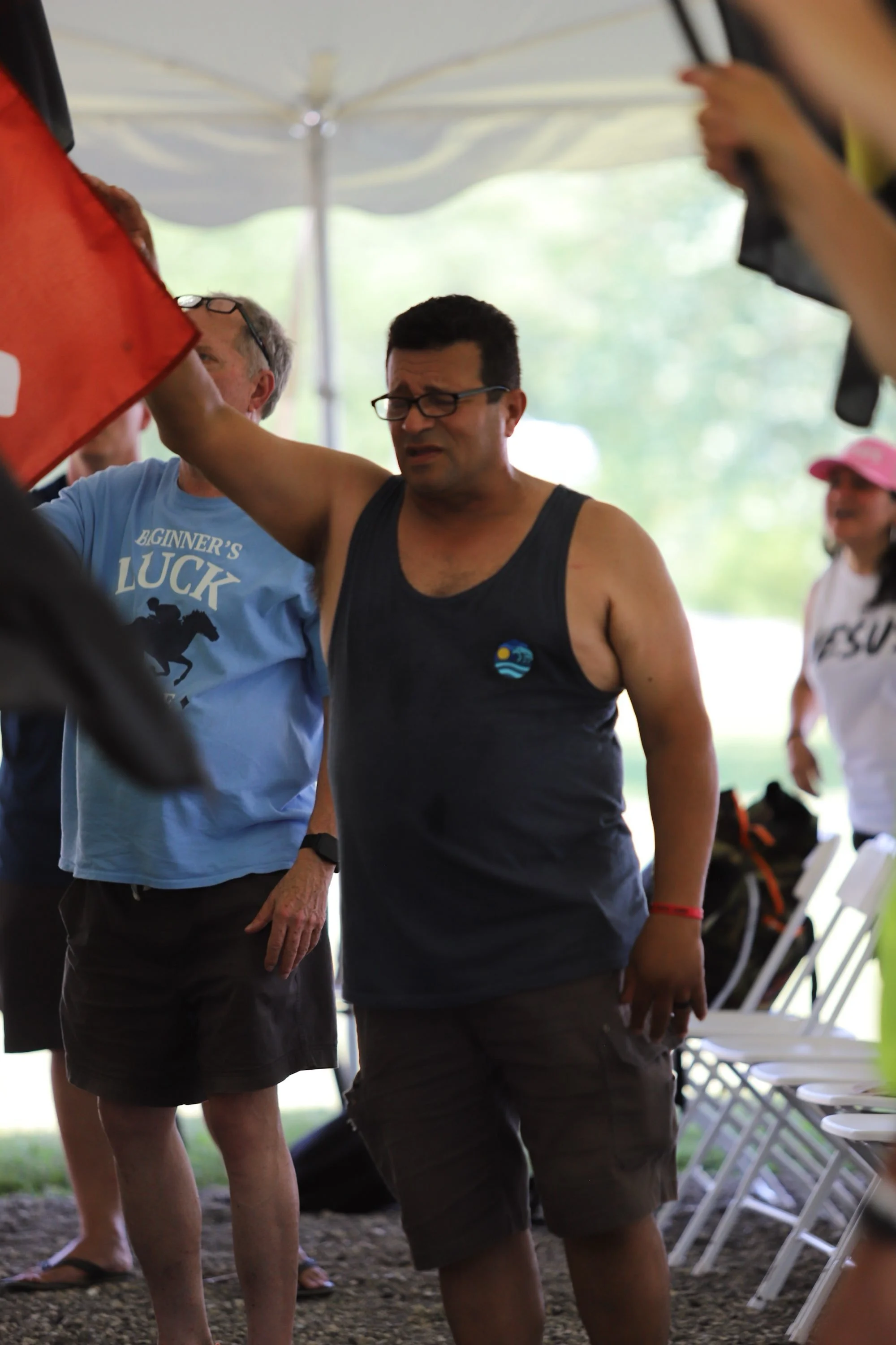 A man in a black tank top and glasses standing outdoors with other people around him, some with chairs, under a canopy or tent, possibly at an event or gathering.
