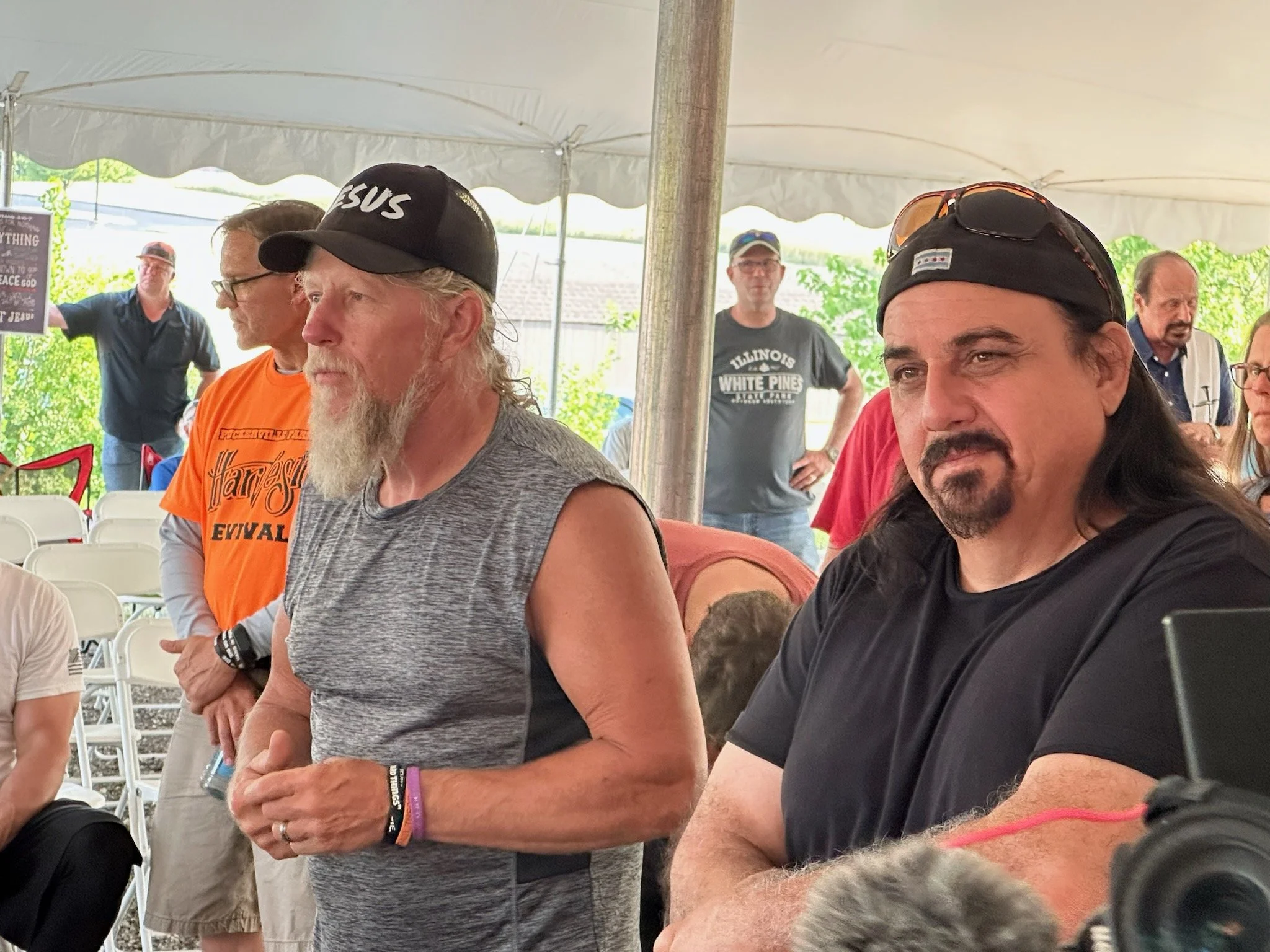 Several men sitting and standing under a white canopy, some wearing baseball caps and casual clothing, engaged in a gathering or event outdoors.