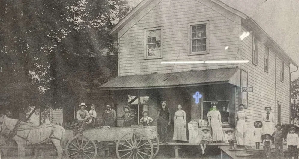 Black and white photo of a two-story wooden house with a group of people, both adults and children, standing outside. The house has a cross sign on the front, indicating it is a church or religious building. There is a horse-drawn cart in front of the house, and the scene appears historical.