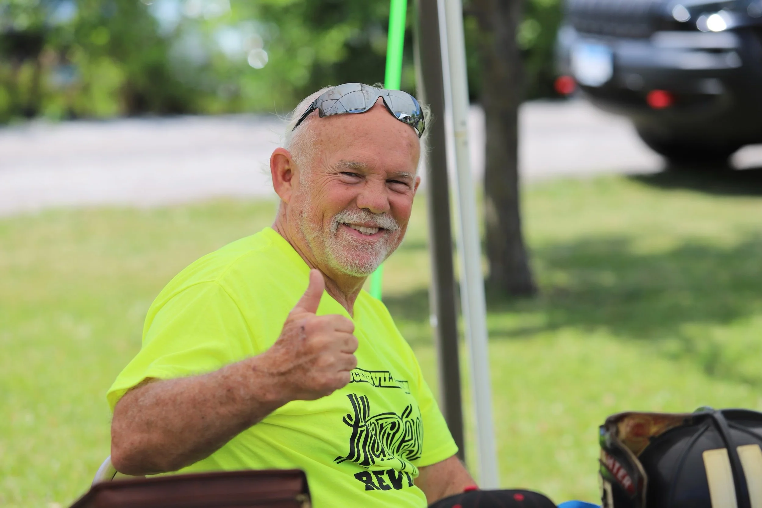 Smiling older man wearing sunglasses and a bright yellow shirt giving a thumbs-up at an outdoor event.