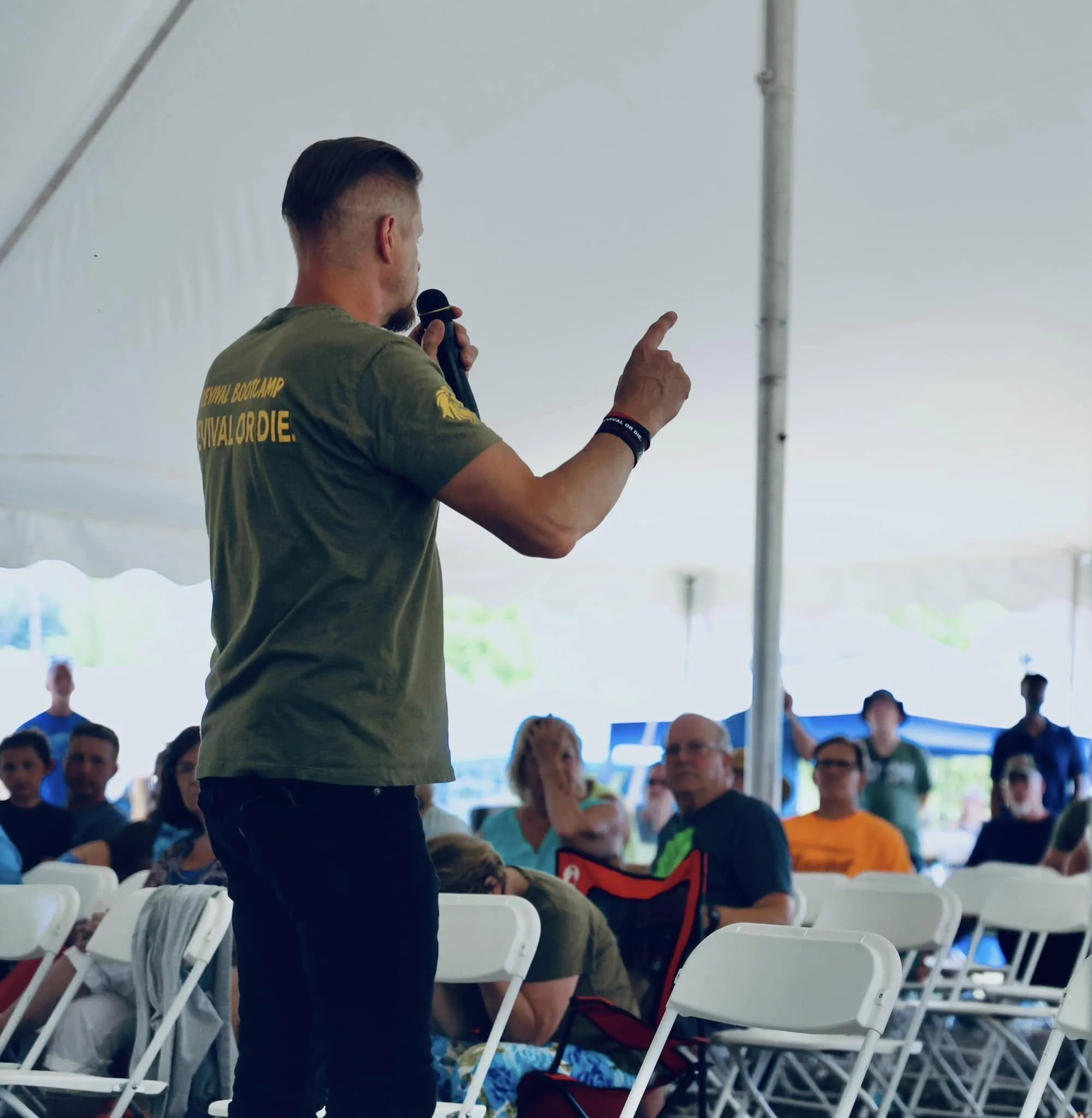 A man with a beard and short hair, wearing a green t-shirt with yellow text and black pants, is speaking into a microphone in front of an audience seated on white chairs under a large tent. The audience appears to be listening attentively.