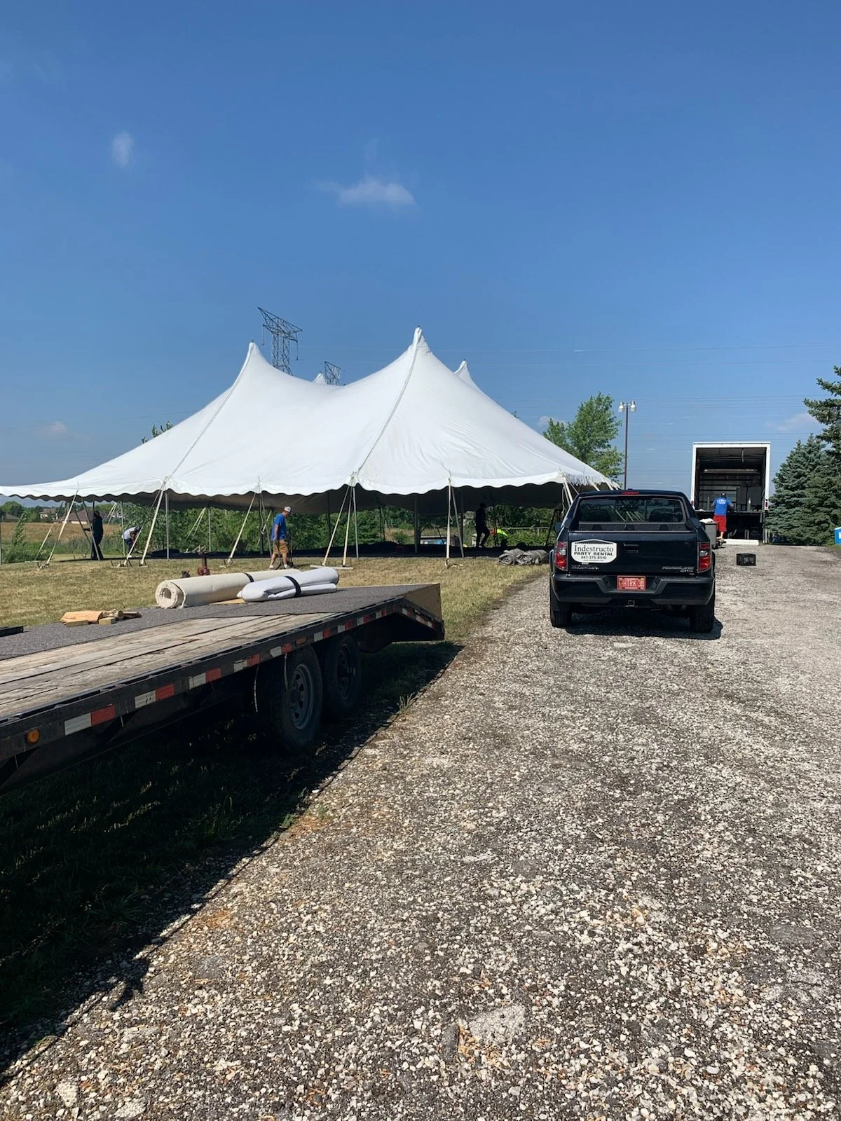 A large white event tent being assembled outdoors with several people working on it. A flatbed trailer with rolled-up material and tools is in the foreground. A black pickup truck and a white moving truck are parked nearby on a gravel path, with tree
