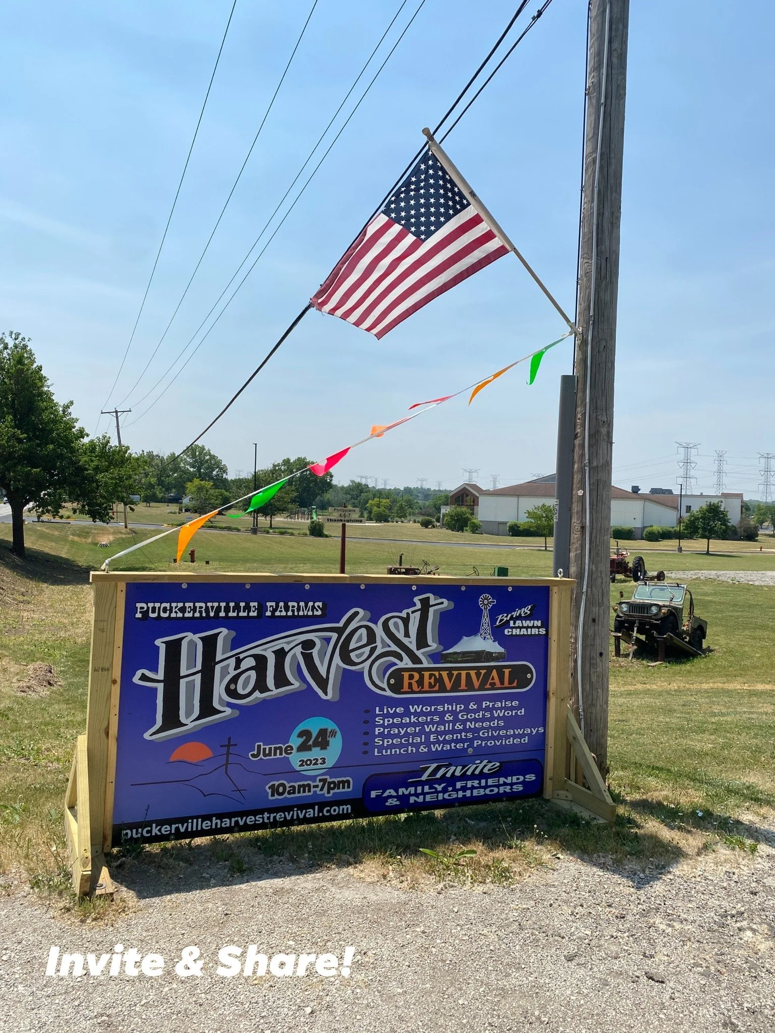A large blue sign for 'Puckerville Farms Harvest Revival' event, featuring details about the date, time, activities, and website, with an American flag and colorful banners overhead, set outdoors on a sunny day.