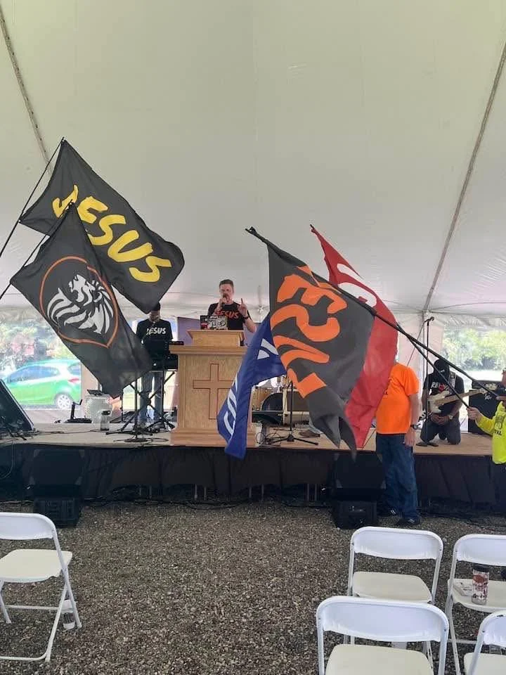 A church service or event taking place inside a large white tent with a stage. There are multiple flags, including a Christian fish symbol and an American flag, held by people in the audience. A woman is speaking at a pulpit, and there is musical equ