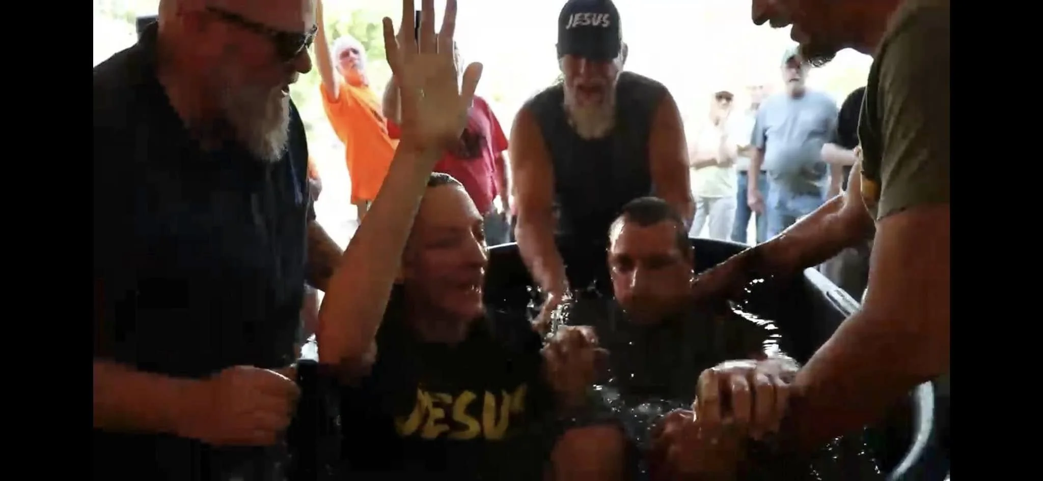 People participating in a baptism ceremony, with one person submerged in water and others assisting and celebrating.