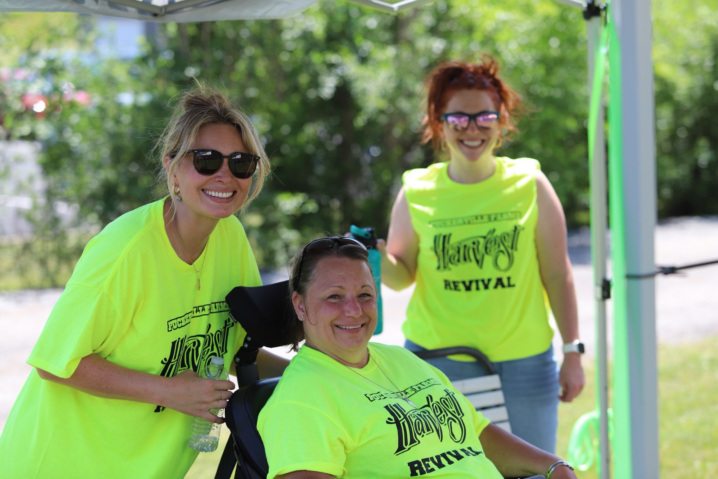 Group of four women wearing bright yellow T-shirts with ‘Harvest Revival’ printed on them, smiling outdoors with green trees in background, some wearing sunglasses, one holding a water bottle.