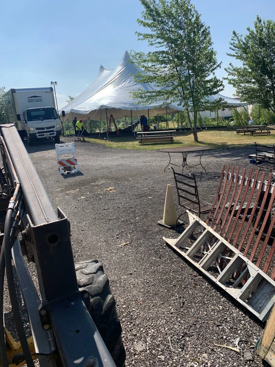 Large white event tent being assembled outdoors with trees and benches around, a truck nearby, and construction equipment and barriers in the foreground.
