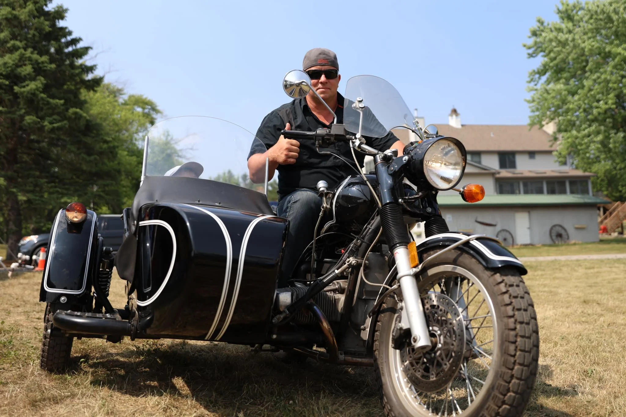 Man wearing sunglasses and a cap riding a black motorcycle with sidecar on a grassy area, with houses and trees in the background.