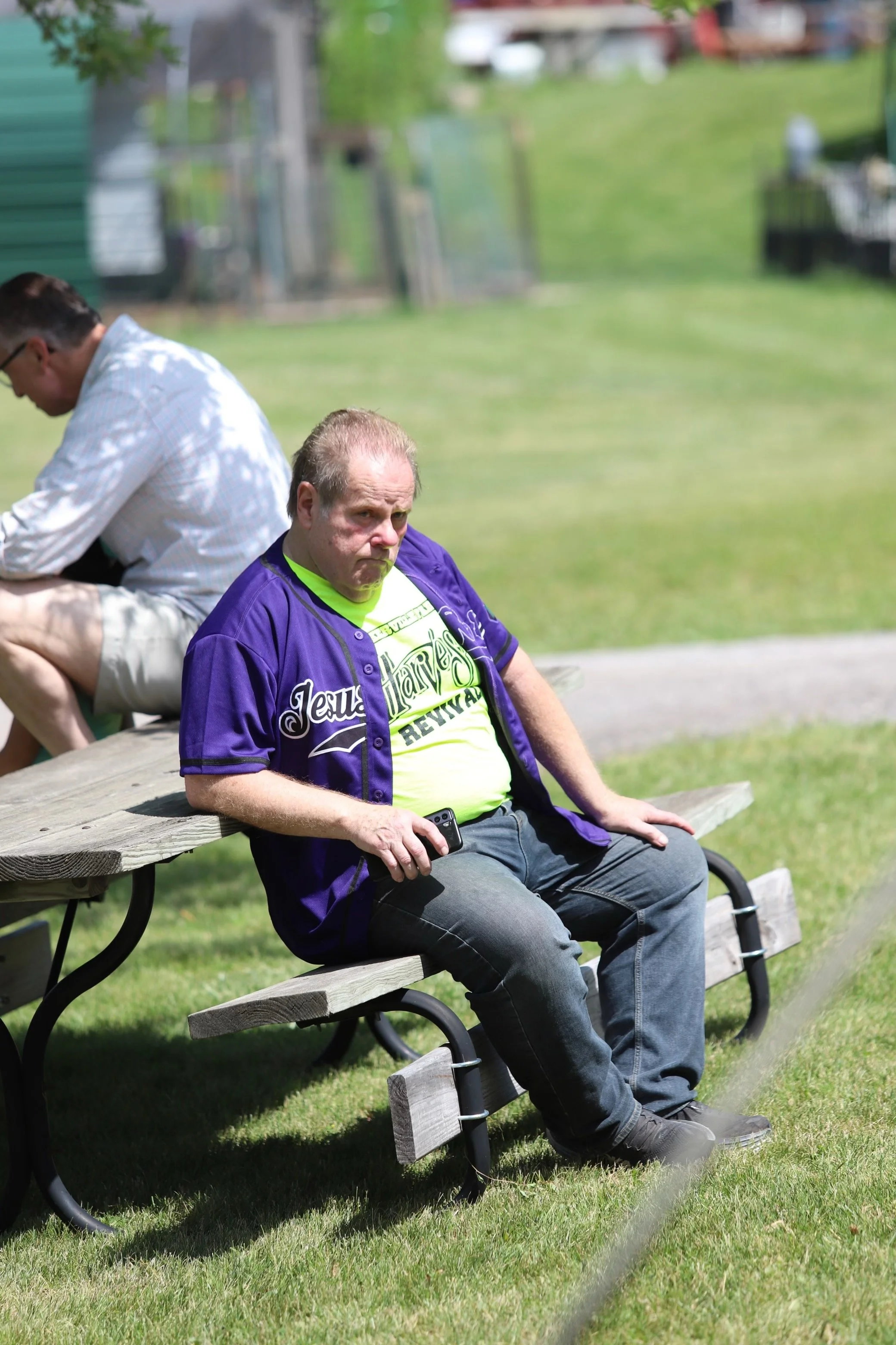 A man with a beard and glasses sitting on a park bench while holding a phone, wearing a colorful sports jersey and a purple jacket. Another man is sitting on the bench with his back to the camera, wearing glasses, a white shirt, and beige shorts. The