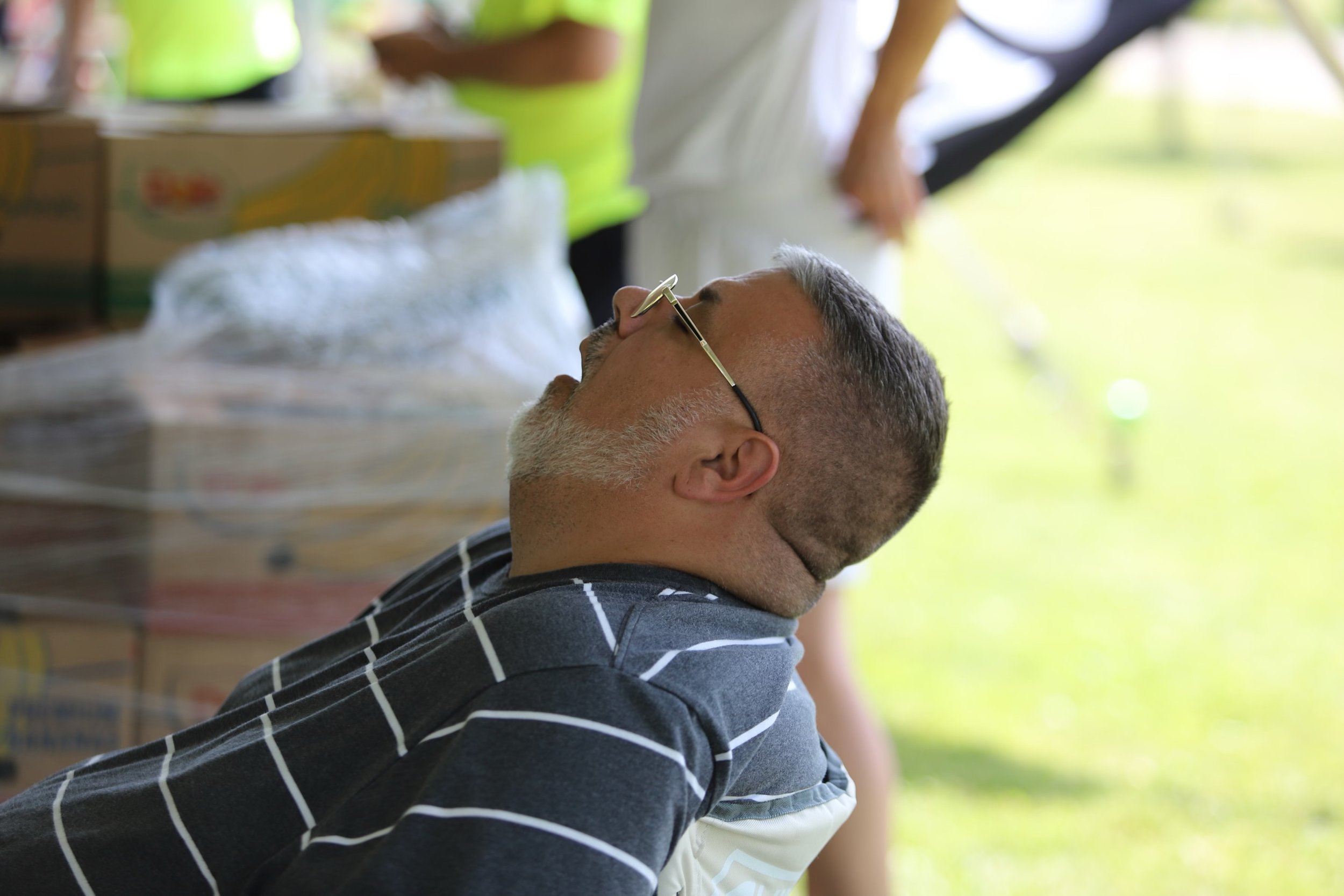 A man with gray hair and glasses, wearing a striped shirt, rests with his eyes closed outdoors during the daytime.