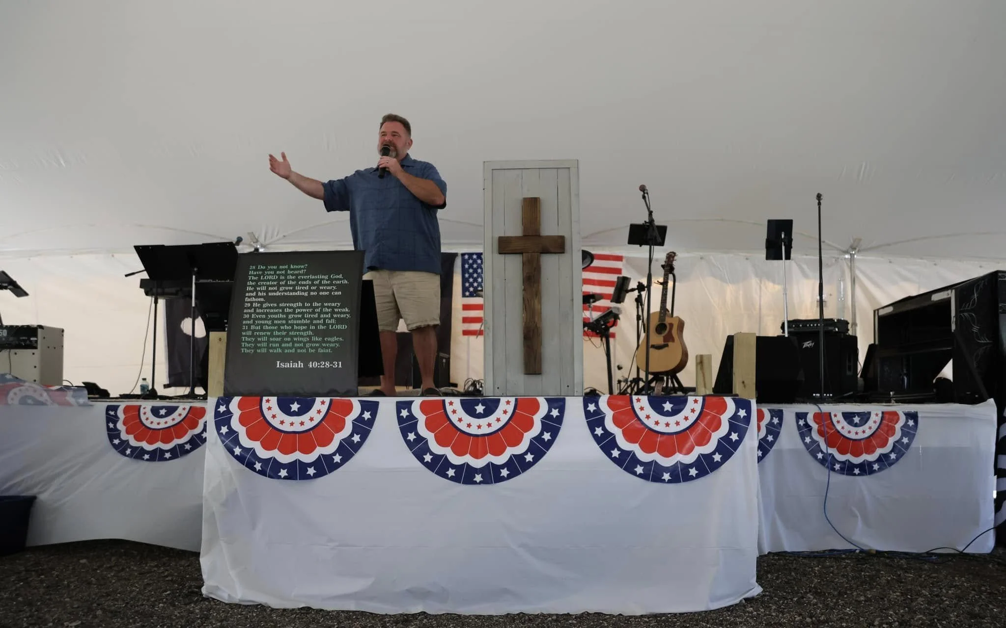 A man speaking on stage at an American patriotic-themed event, with a cross on the stage, American flags, and musical instruments including guitars, set up inside a tent.