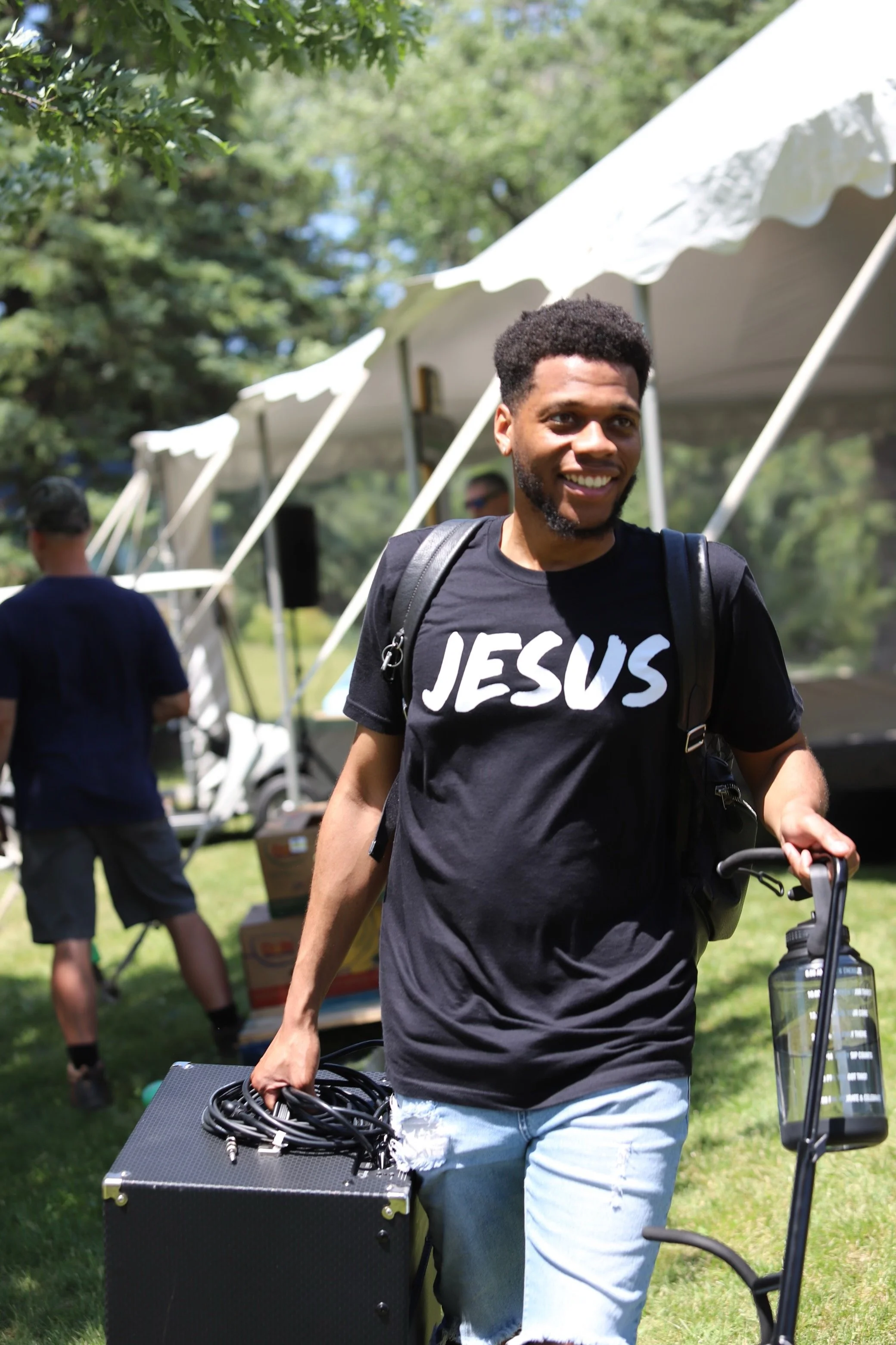 A young man with curly hair and a beard smiling while walking outdoors during daytime, carrying a black case with coiled cables in one hand and a water bottle in the other. He is wearing a black T-shirt with white text that reads 'JESUS' and light bl
