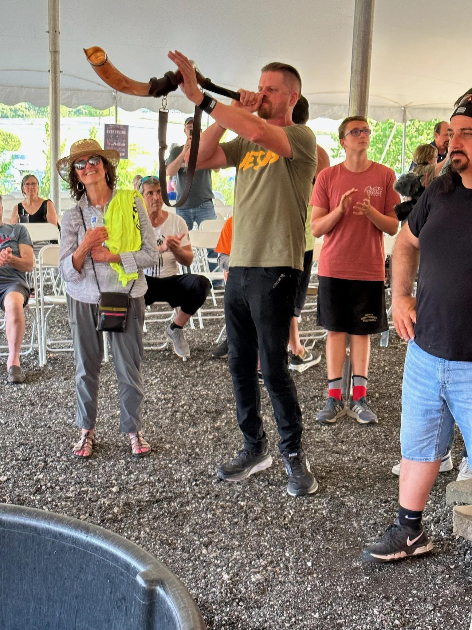 A man playing a wooden wind instrument at an outdoor event under a tent, with several seated and standing spectators watching.