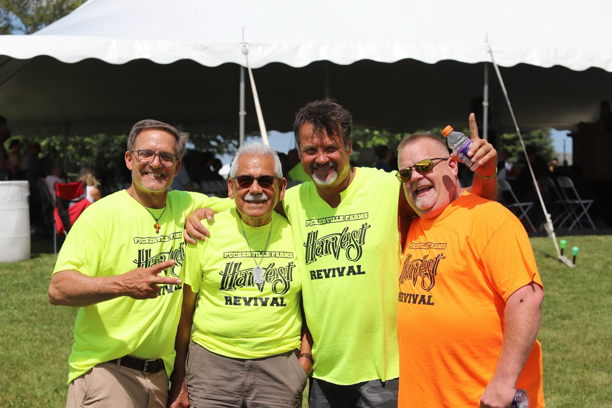 Four men smiling and posing outdoors under a tent, wearing bright yellow and orange shirts with the words "Harvest Revival" printed on them, at a daytime event.