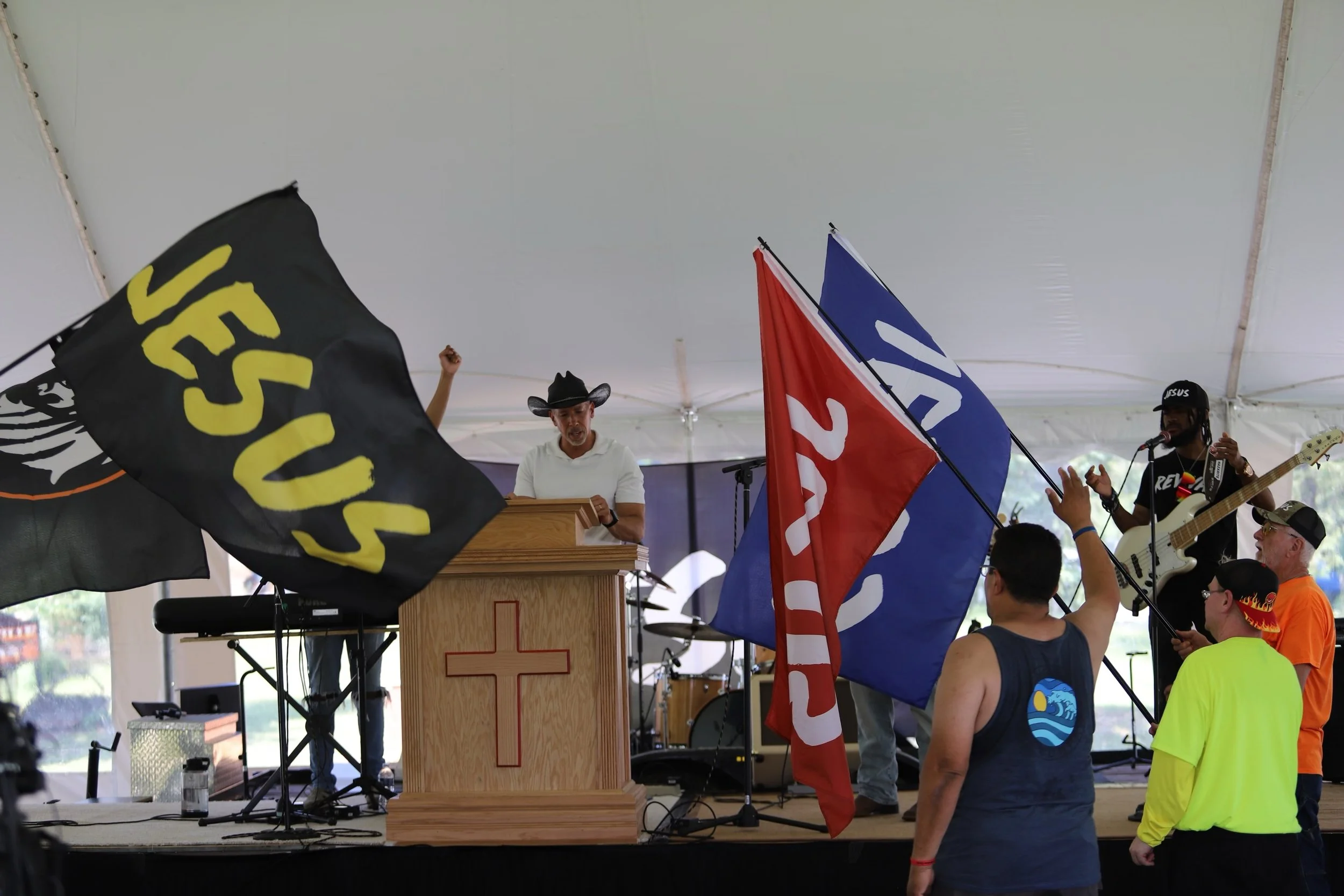 A group of people attending a religious event under a tent, with two men playing guitars, one woman speaking at a podium, and flags of different colors and designs in the foreground.