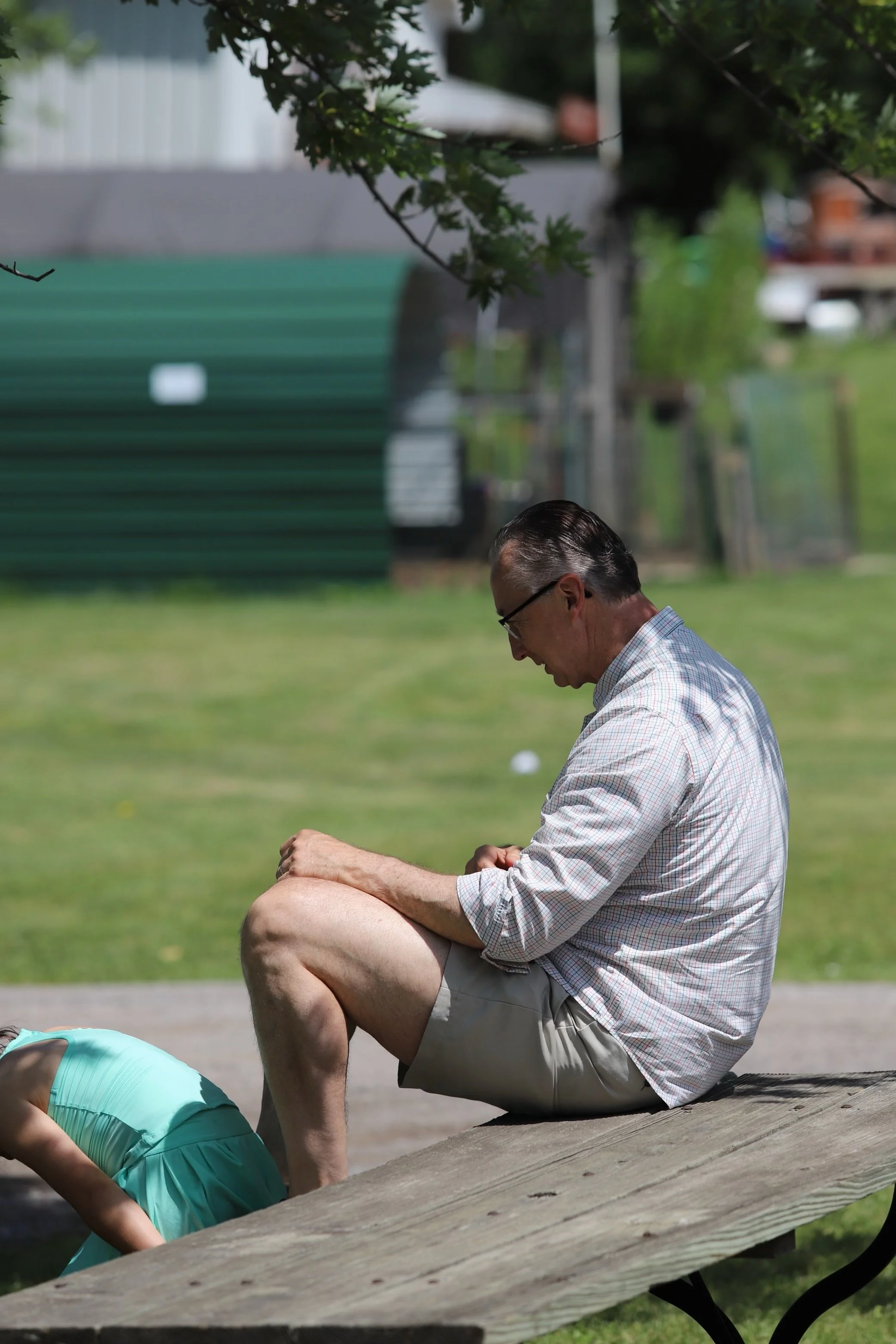 An older man sitting outdoors on a wooden bench, wearing glasses, a checkered shirt, and shorts, while a young girl in a light blue dress bends over in front of him.
