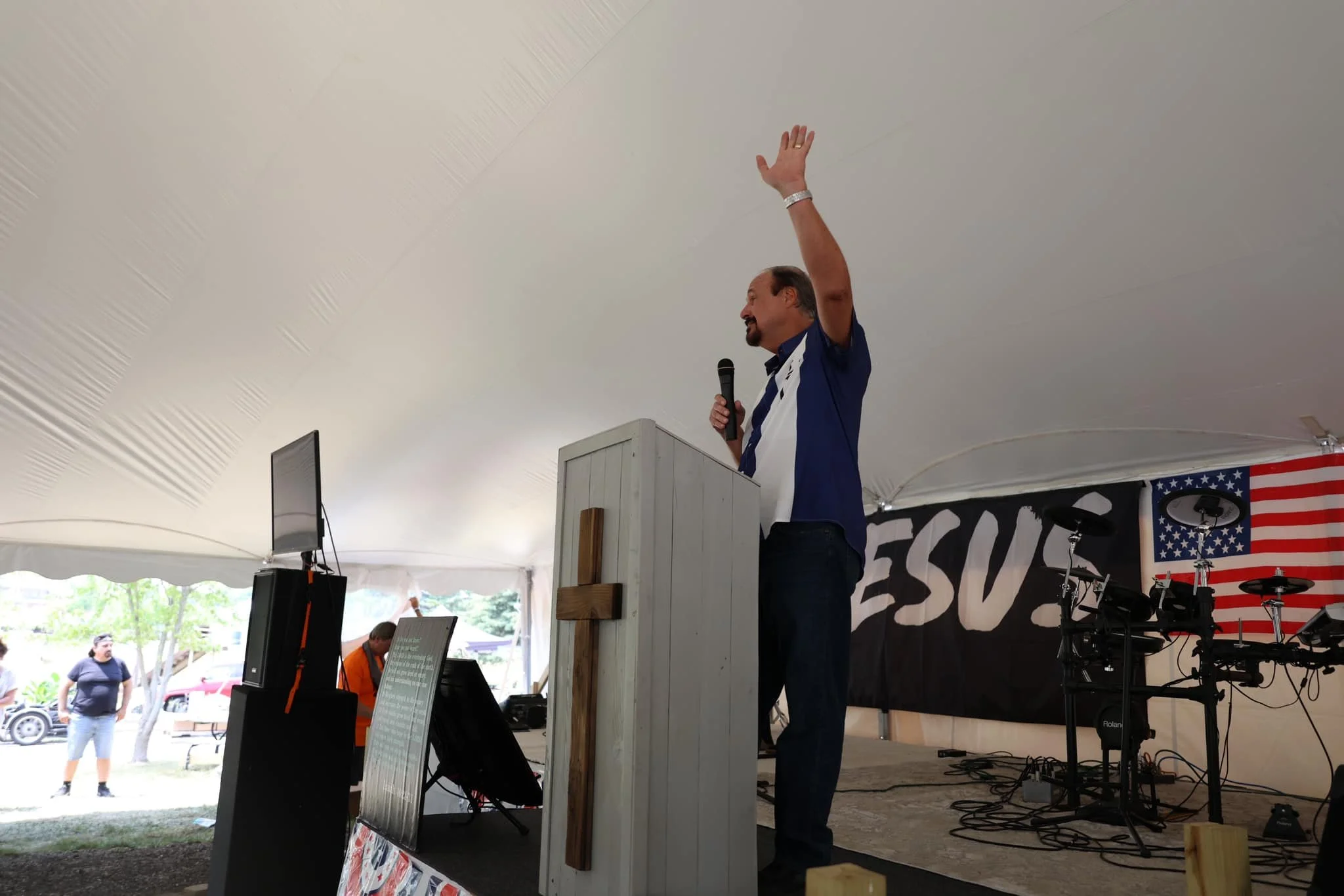 A man is speaking at a podium during a patriotic event inside a tent, with an American flag and percussive set behind him. People are standing outside in the background.