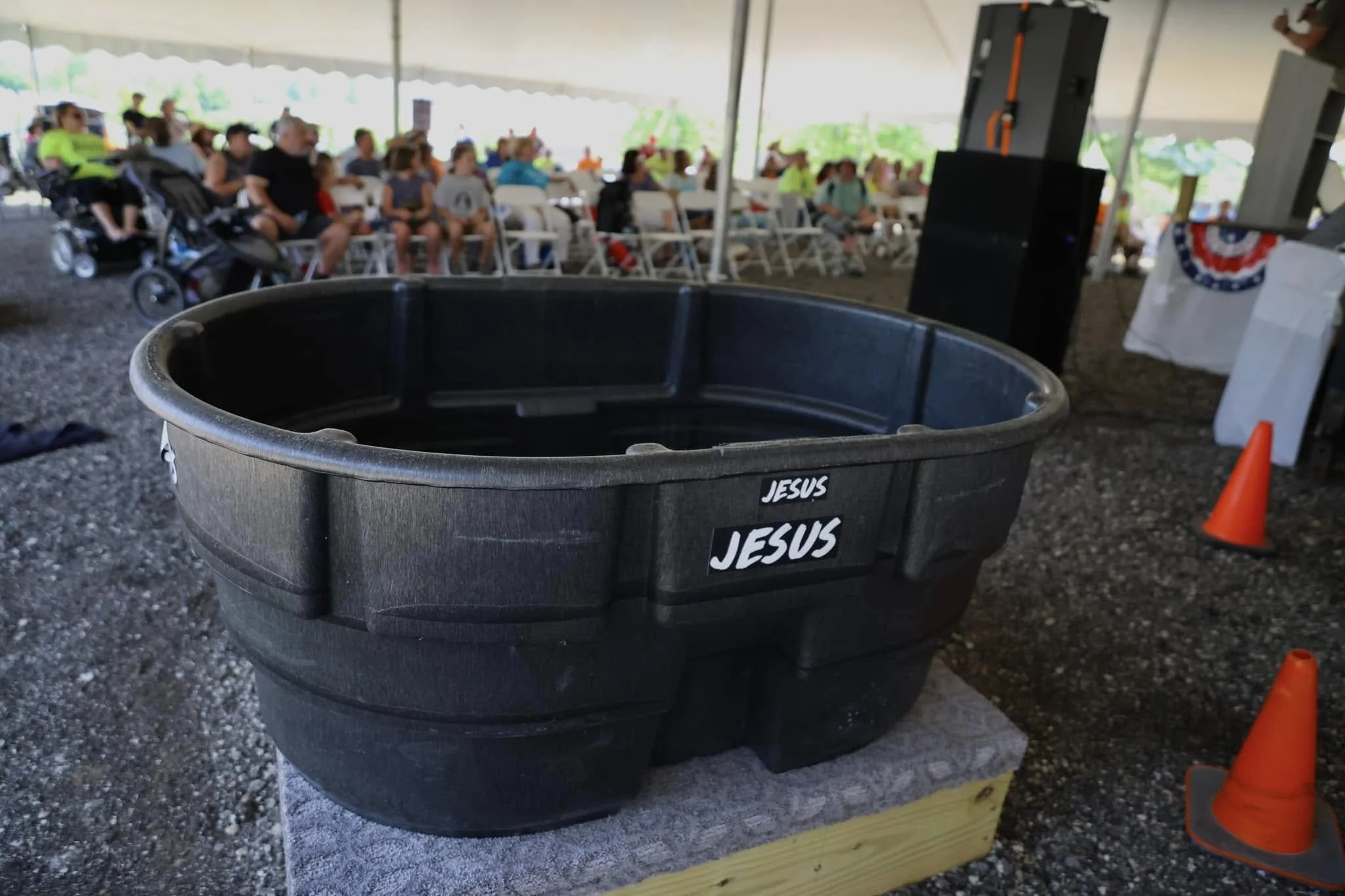 A large black trash can with the word 'JESUS' written multiple times on it, placed on a gravel floor inside a large tent. In the background, there is a crowd of people seated on chairs, some wearing bright colors, with a blurred outdoor setting visib