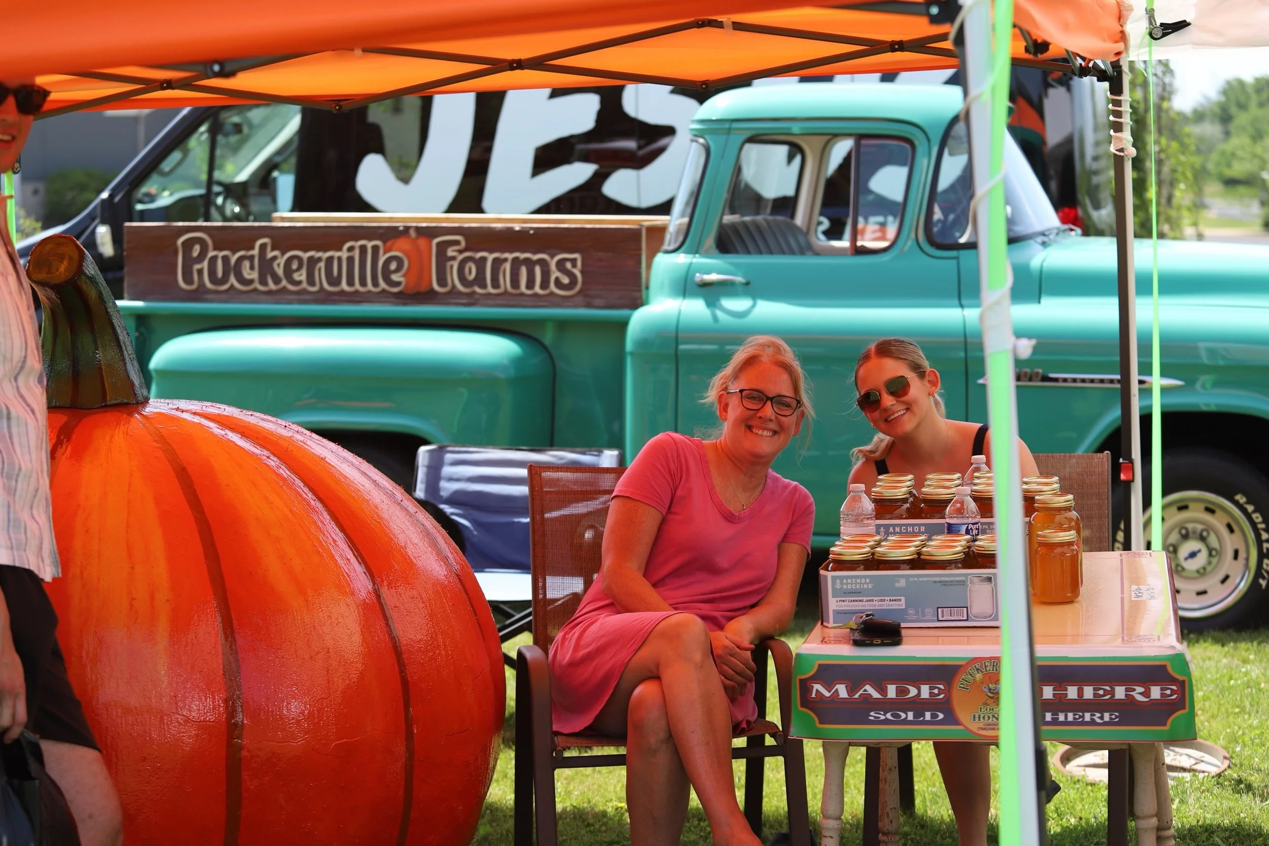 Two women sitting at a table outdoors at a farm stand, smiling, with jars of honey on the table. Behind them is a vintage teal truck with a sign that reads 'Pockerille Farms.'