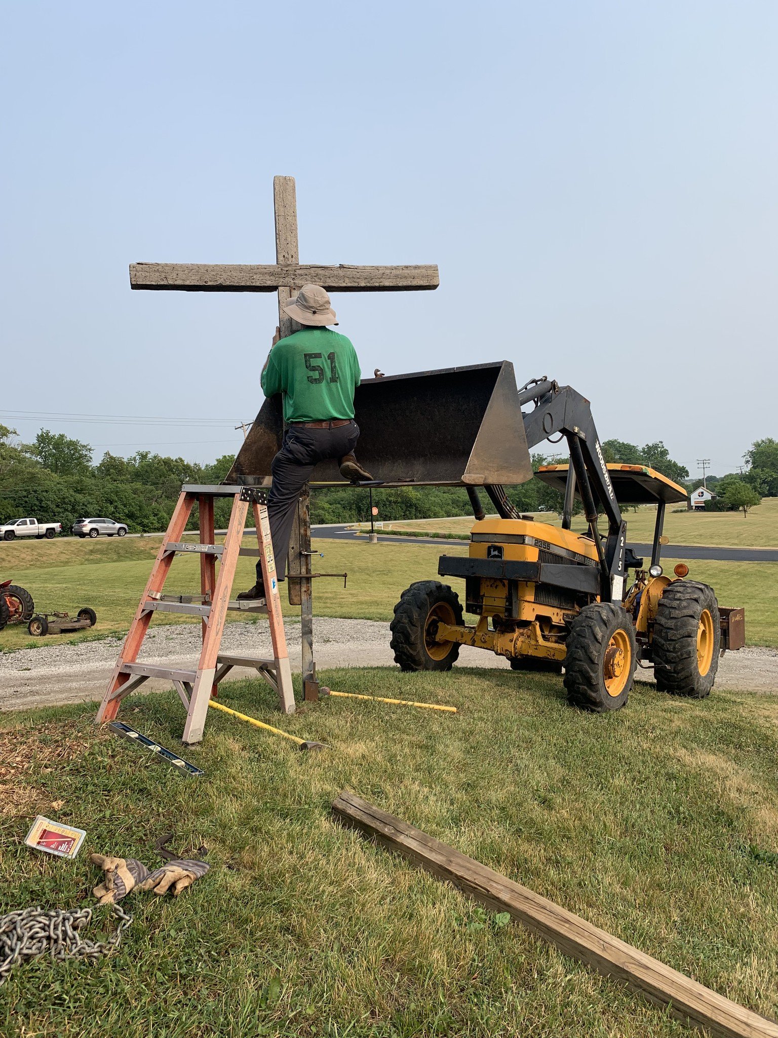A man in a green shirt and tan hat kneeling on a ladder, securing a wooden cross to a metal post. The scene is outdoors with a yellow tractor, parked cars, and trees in the background.