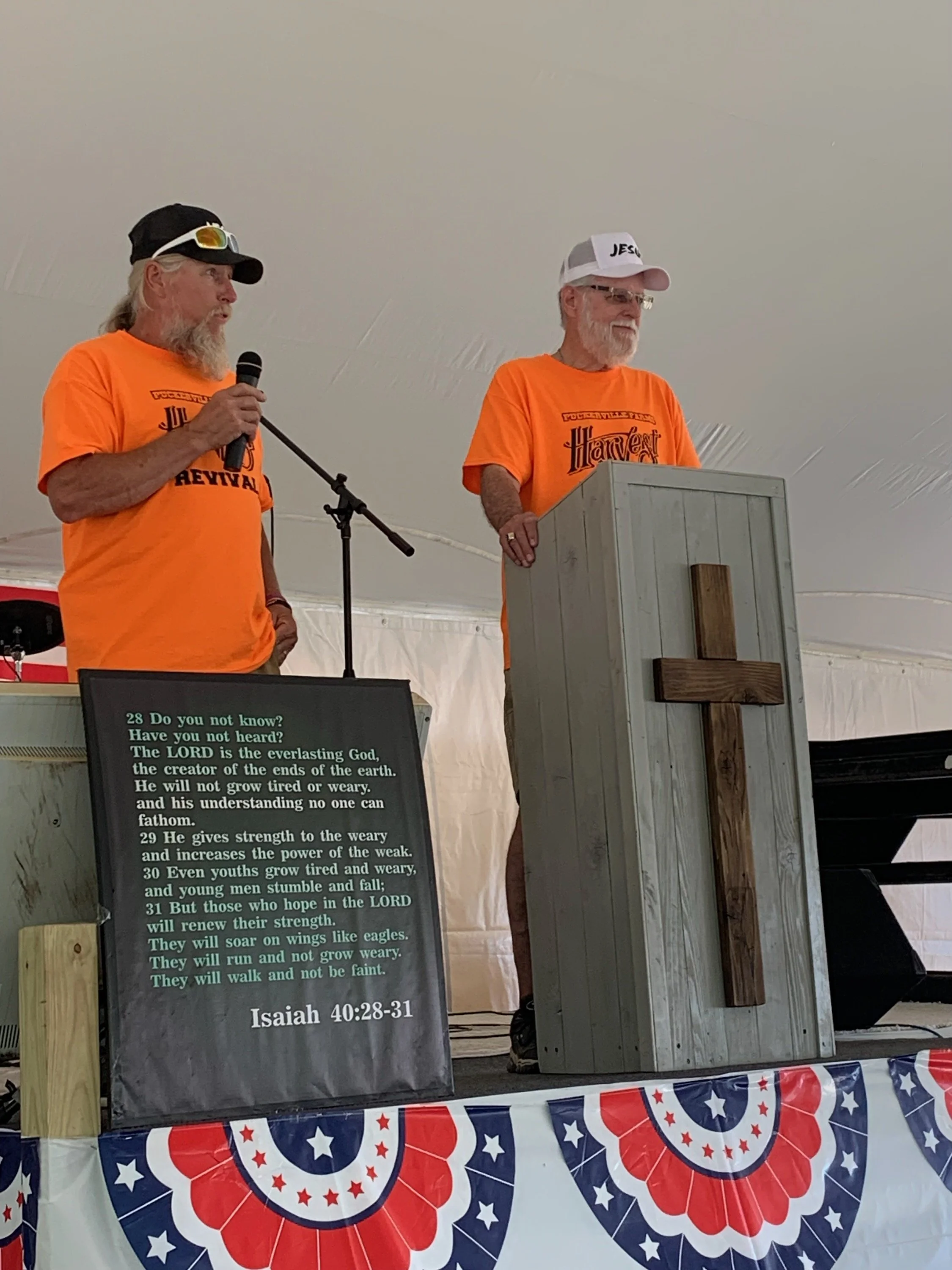 Two elderly men with white hair and beards, wearing orange shirts with 'Harvest Revival' printed on them, stand behind a podium. One man is holding a microphone and speaking, while the other stands beside him. Both are under a white tent decorated wi