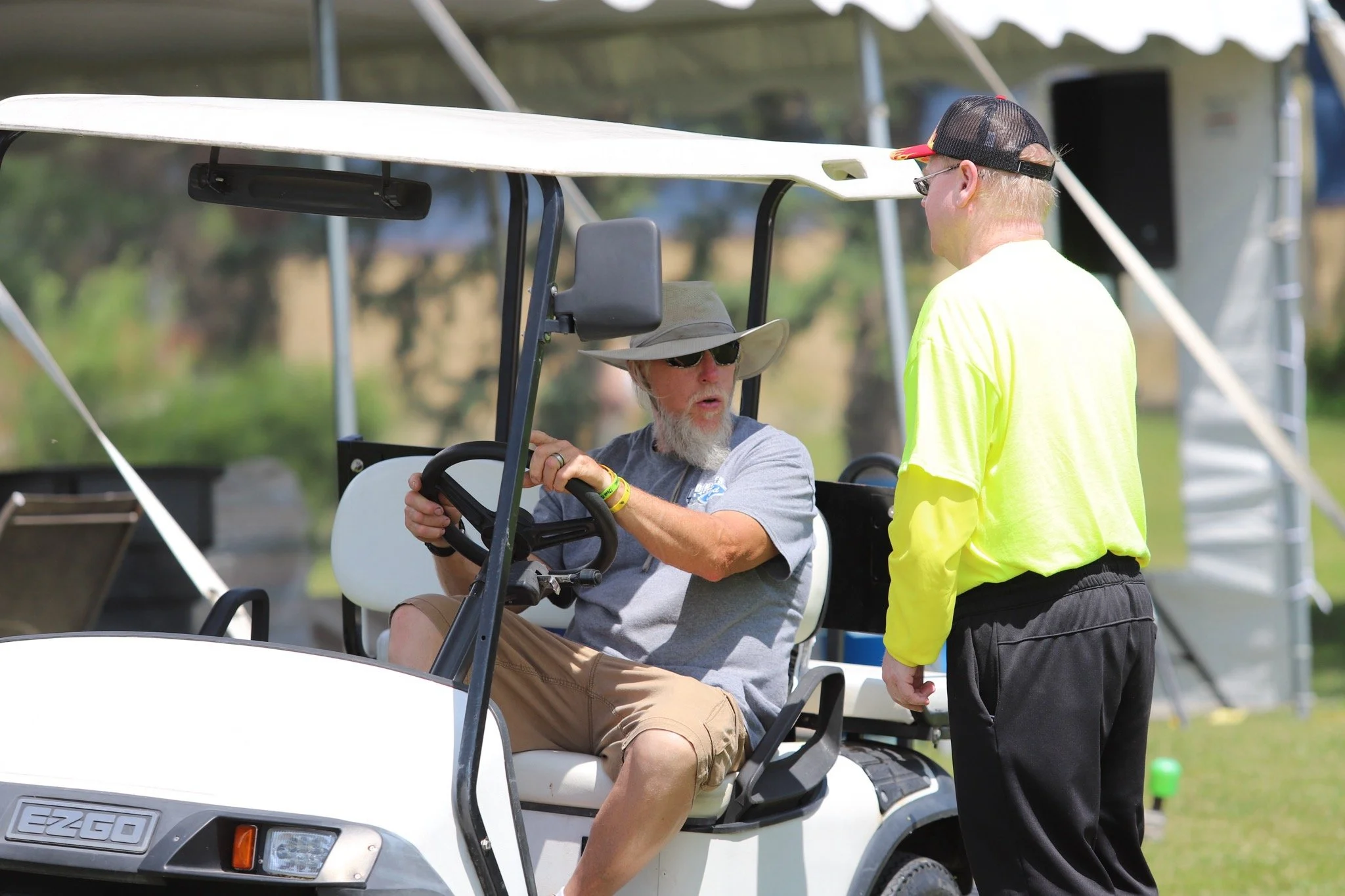 A man with a beard, wearing sunglasses, a gray t-shirt, and khaki shorts, sits in a white golf cart. He is holding the steering wheel and looking at a person standing next to him. The person standing is a woman with blonde hair, wearing a neon yellow