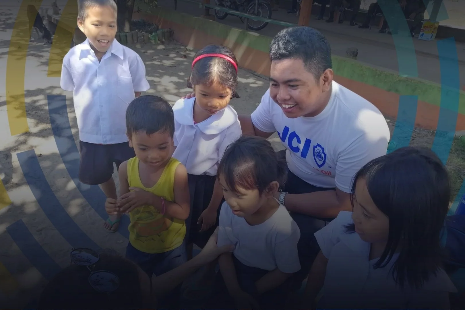 A group of young children gathered outdoors around a smiling man wearing a white T-shirt, engaged in an activity with small containers. The children are attentively watching and listening, appearing curious and interested.