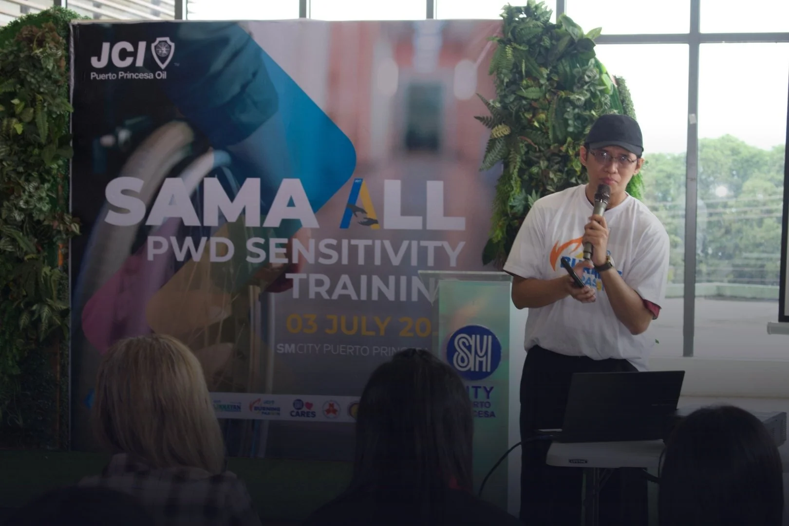 A woman wearing a black cap and glasses presenting at a workshop or seminar, standing beside a presentation podium, with a large colorful banner behind her that reads 'SAMALL PWD Sensitivity Training' with the date '03 July 20' and the location 'SM City Puerto Princesa'.