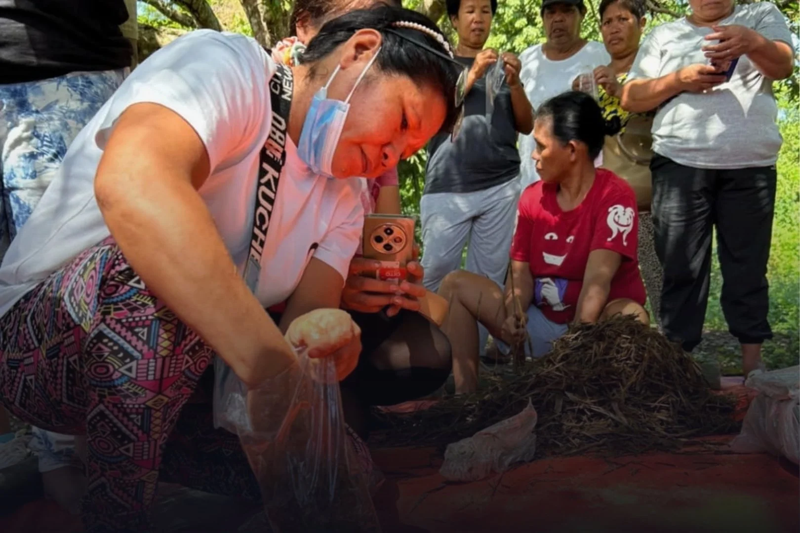 A group of people gathered outdoors around a woman with long black hair, who is sitting on the ground and appears to be involved in a traditional activity. The woman is wearing a red shirt, and some of the people surrounding her look on with interest. One person in the foreground is taking a photo or video with a smartphone. The setting appears to be a wooded area with sunlight filtering through the trees.