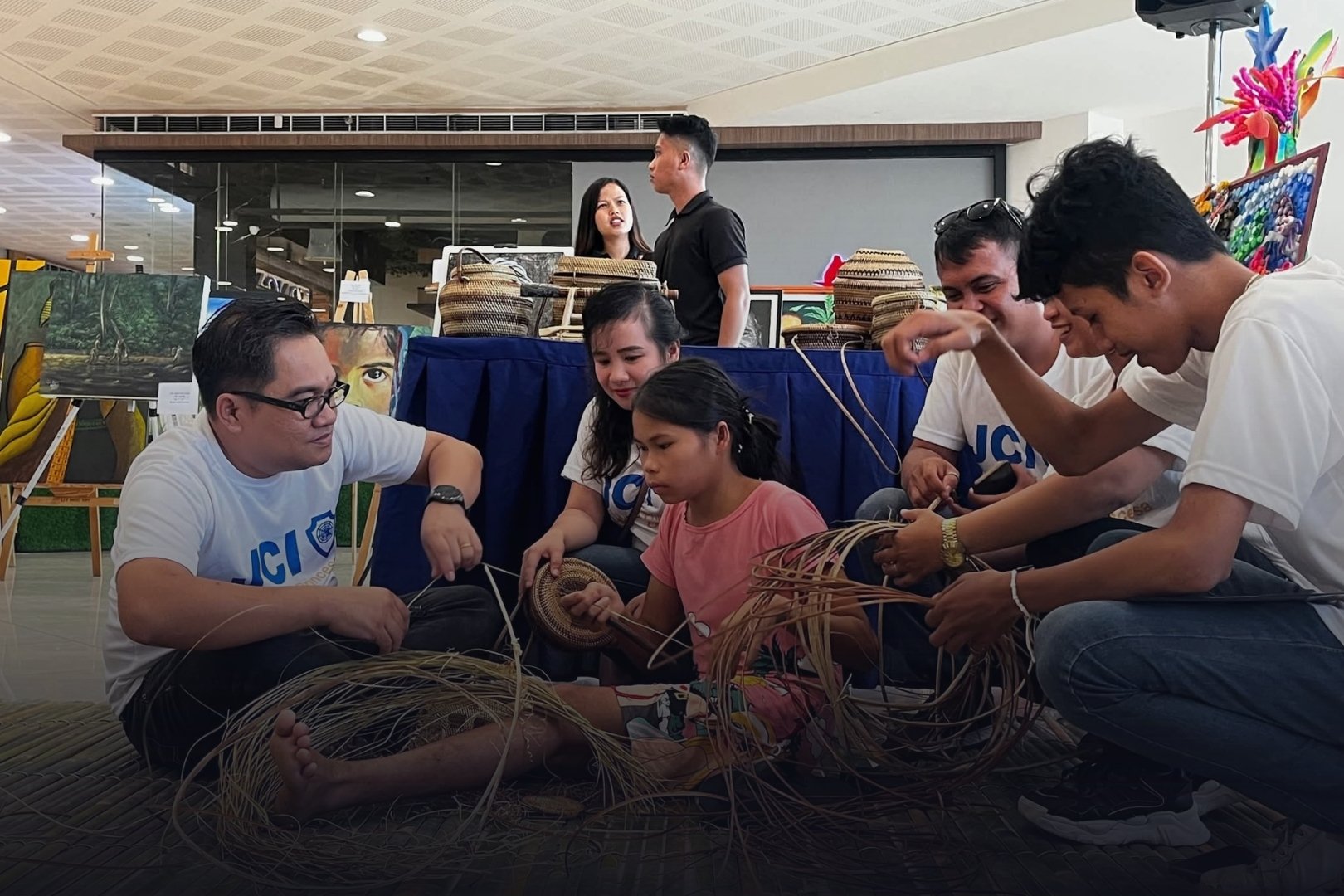 Group of people sitting on a mat, making a craft with natural materials, with an art display and vendor booth in the background.