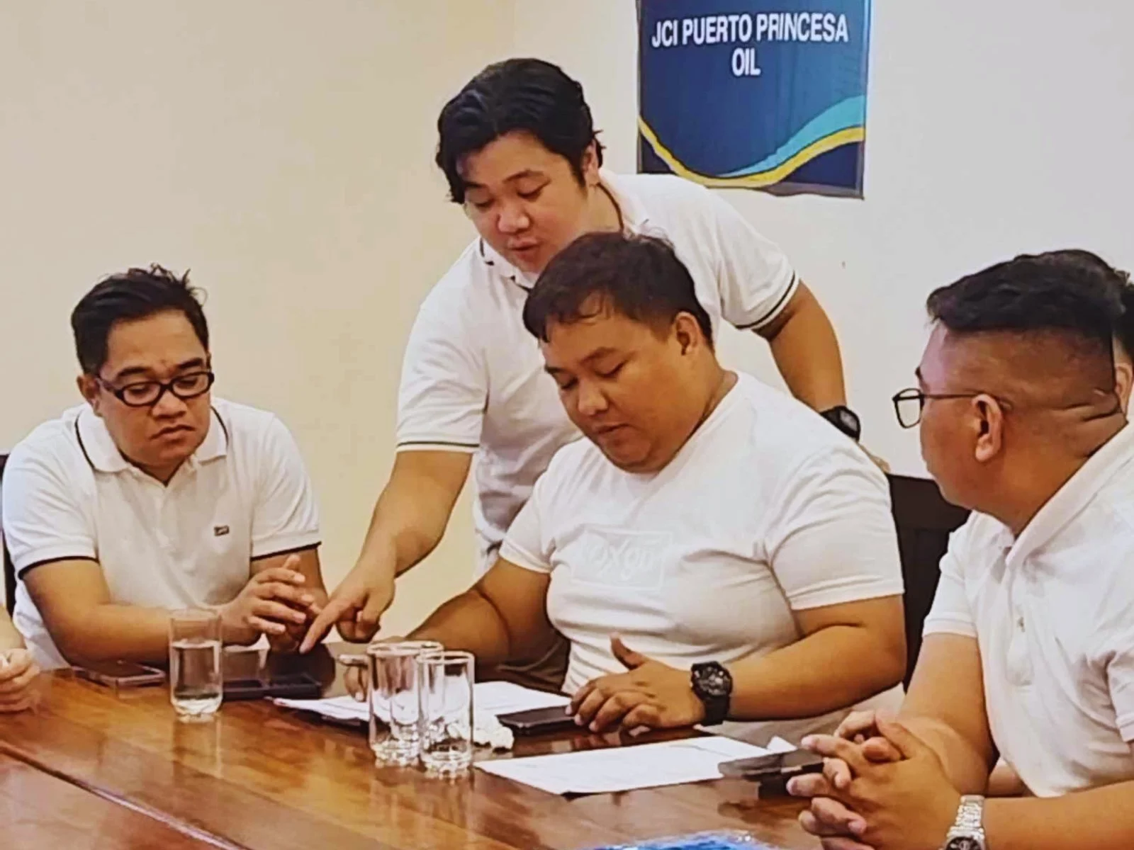 Four men in white shirts sitting and standing around a table with glasses of water, looking at papers and a smartphone, in a room with a sign that says 'JCI Puerto Princesa Oil' on the wall.