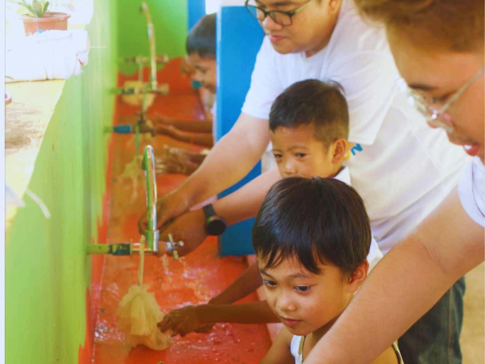 Children washing their hands at a sink with the help of adults in a colorful classroom or community center.