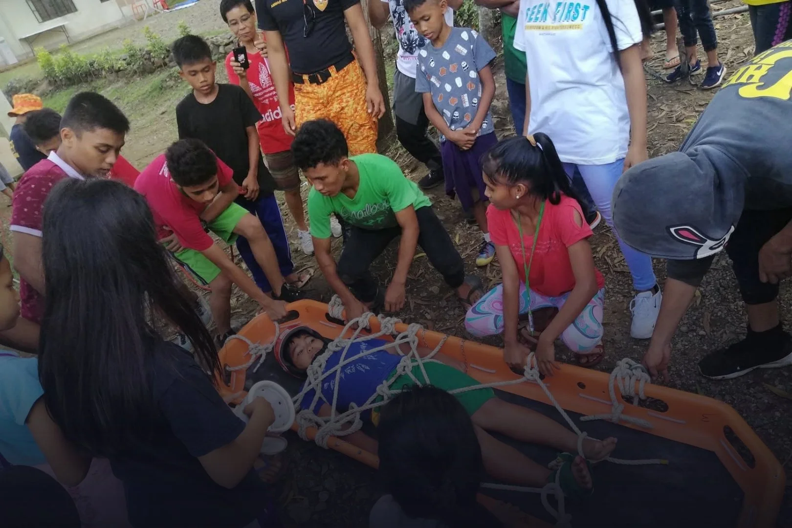 Children and adults gathered around a child on a stretcher with ropes, participating in a rescue drill outdoors.
