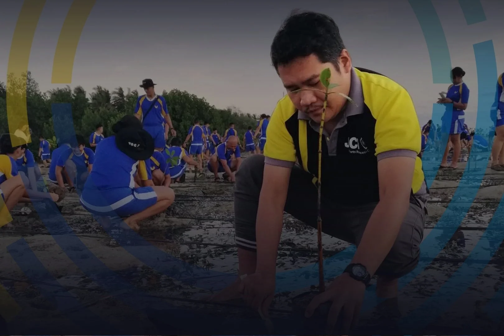 Man planting a tree in a muddy field surrounded by children and people, some in blue and yellow uniforms, during a tree planting event.