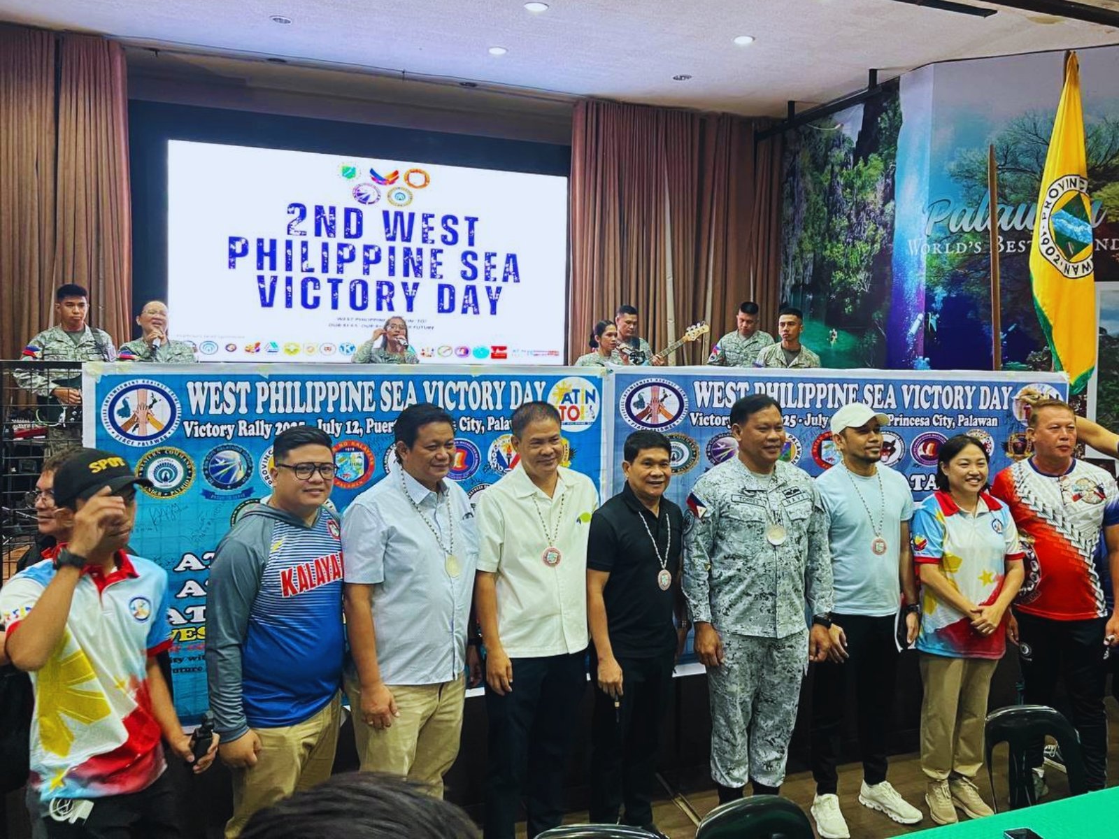 Group of people on stage attending the 2nd West Philippine Sea Victory Day celebration, with a large banner displaying the event's name and date, accompanied by a band performing in the background.