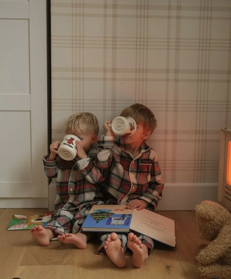 Two young boys in matching plaid pajamas sitting on the floor, drinking from mugs, with a children's book open in front of them, beside a stuffed animal, next to a space heater.